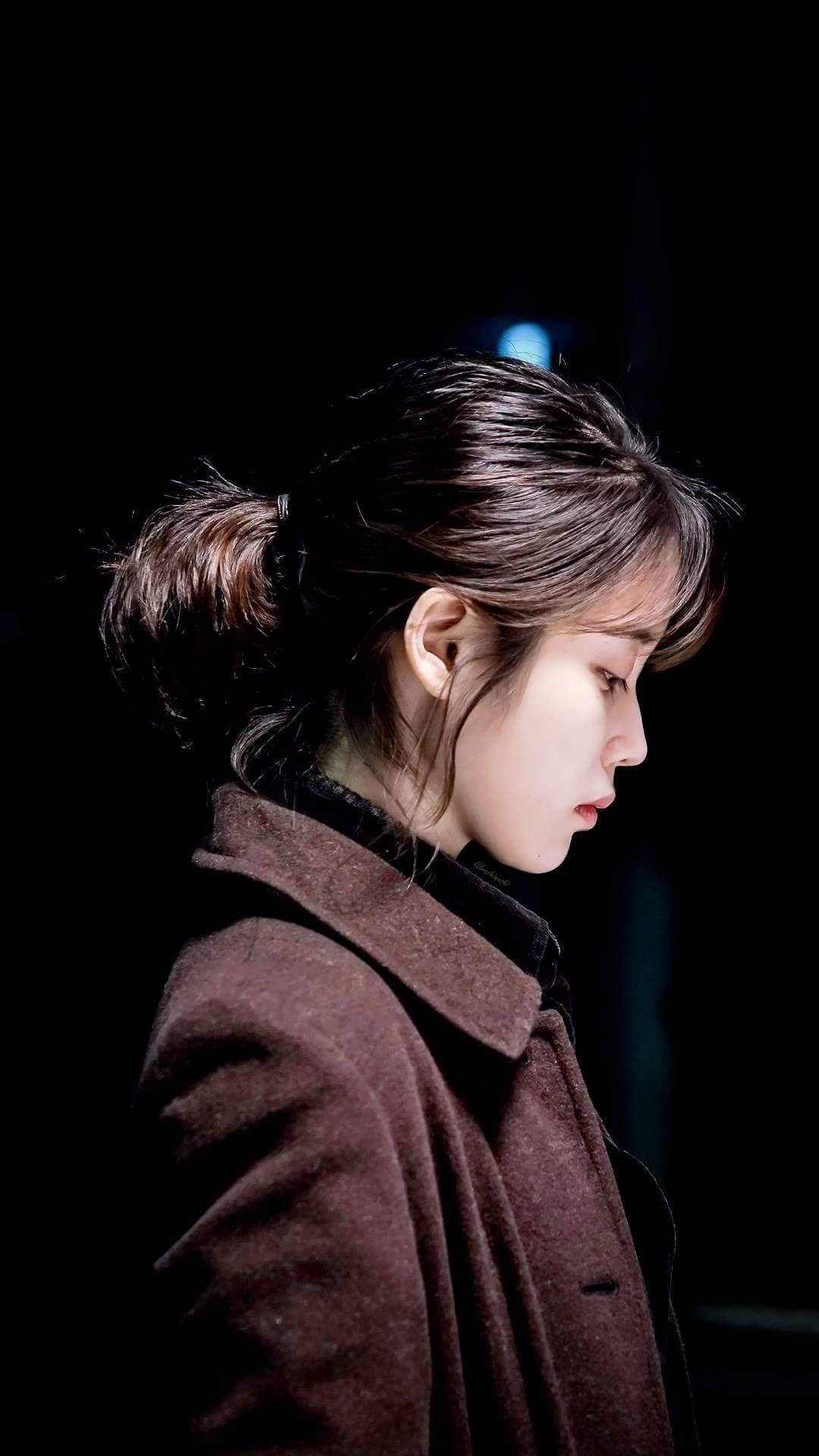 iu phone 桌布 壁紙 — 一位身穿棕色外套的女性站在陰影中，柔和的光線凸顯她的側面和深色頭髮。
