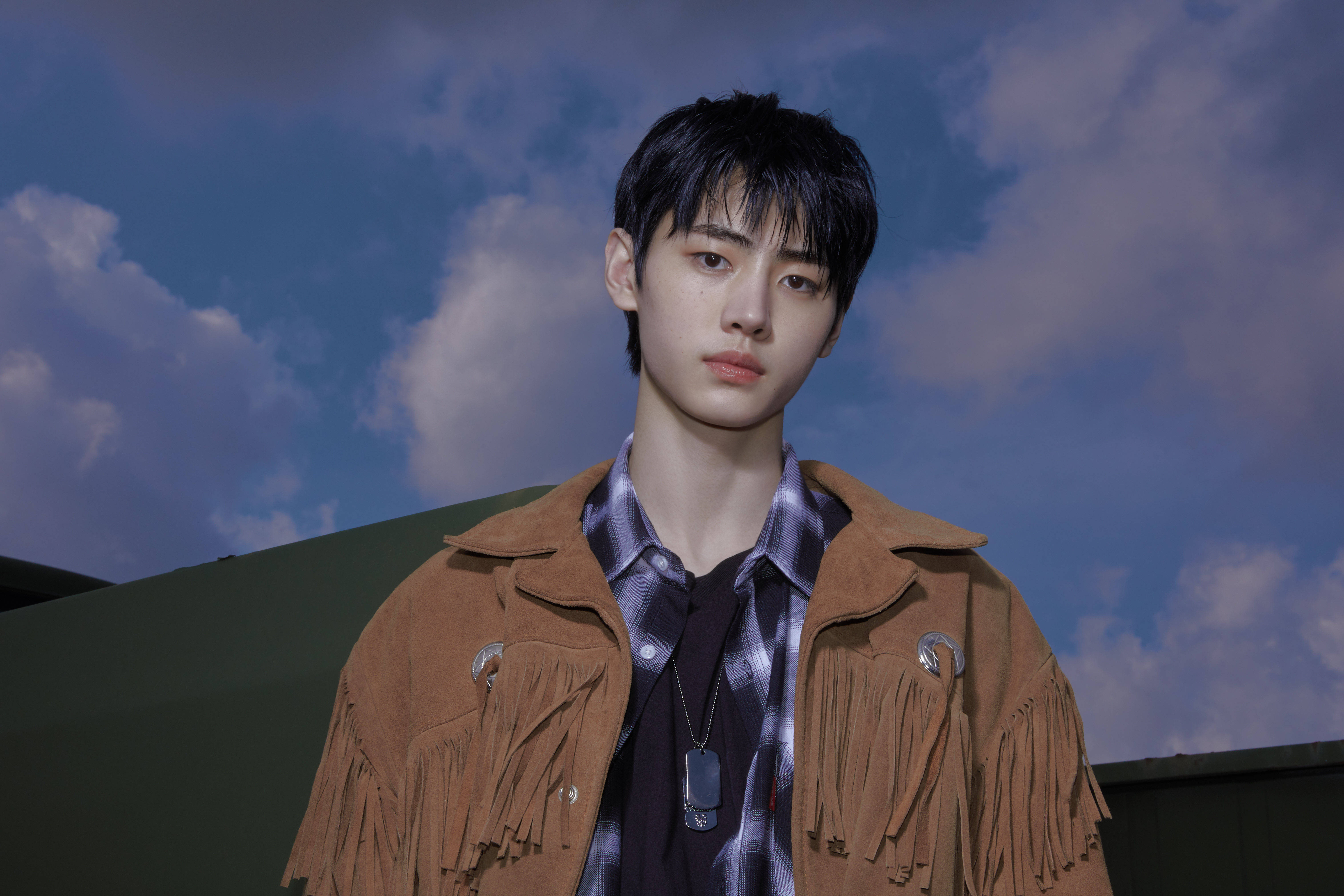 Enhypen Aesthetic 桌布 壁紙 — 一位身穿棕色流蘇麂皮外套的年輕男子，站在戲劇性的藍天白雲背景下。