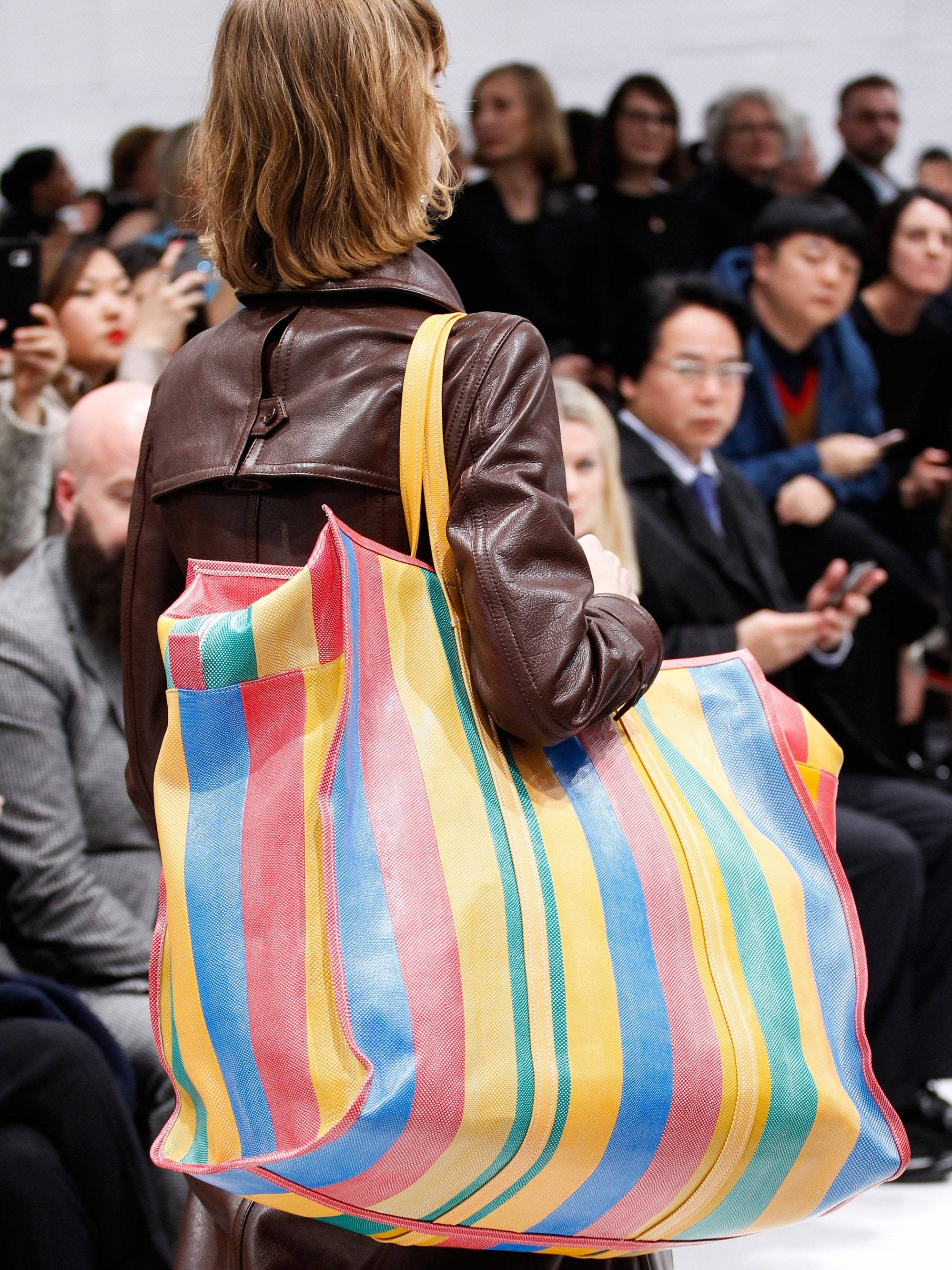 Balenciaga 桌布 壁紙 — 一位身穿棕色皮夾克的女性，在觀眾注視下，拿著一個色彩繽紛的條紋大手提袋走過伸展台。