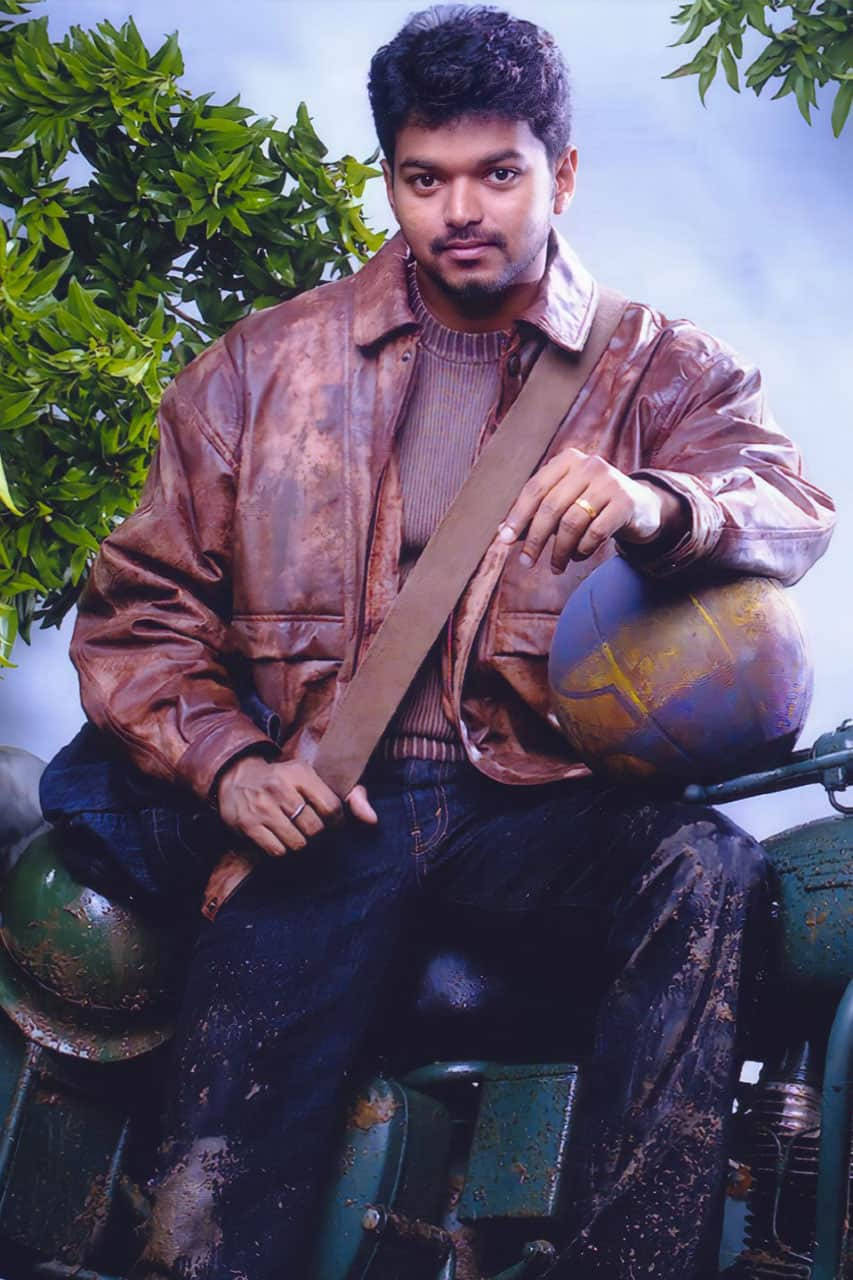 Bollywood HD 桌布 壁紙 — 一位身穿棕色皮夾克的年輕男子自信地坐在一輛復古摩托車上，手持一個地球儀，背景是綠色樹葉和陰天。