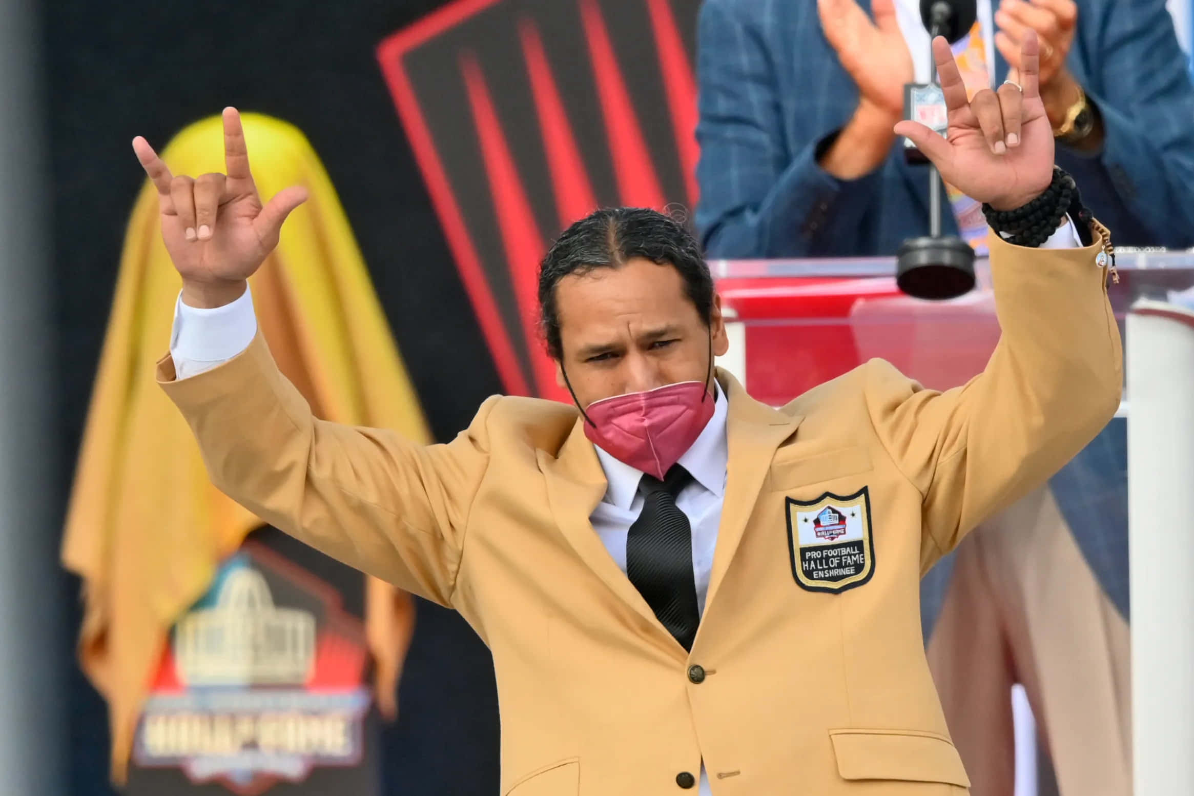 Troy Polamalu 桌布 壁紙 — 一位身穿棕褐色名人堂外套和粉紅色口罩的足球傳奇人物在體育場儀式上做出搖滾手勢。