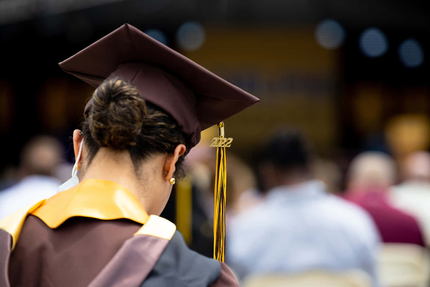 Graduation Ceremony 桌布 壁紙 — 一位身穿棗紅色畢業袍、頭戴2022金色流蘇方帽的畢業生，低頭看著背景模糊的典禮人群。