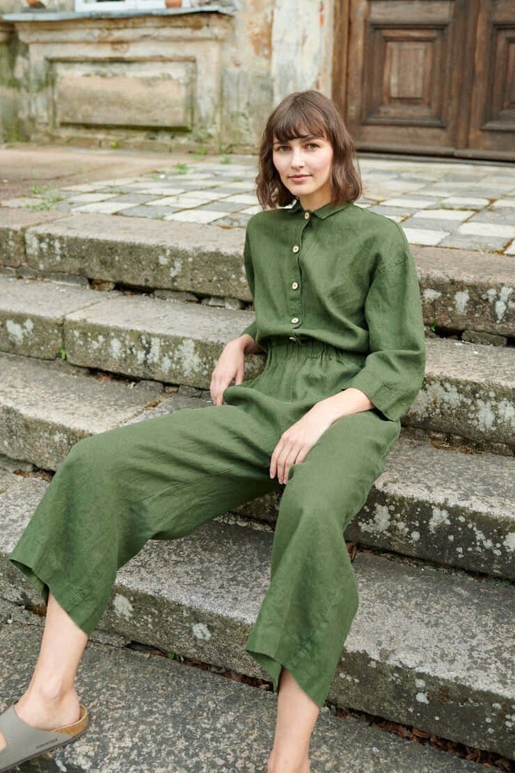 Green Fashion 桌布 壁紙 — 一位身穿橄欖綠亞麻連身褲的女性，平靜地坐在風化的石階上，背景是質樸的建築外牆。