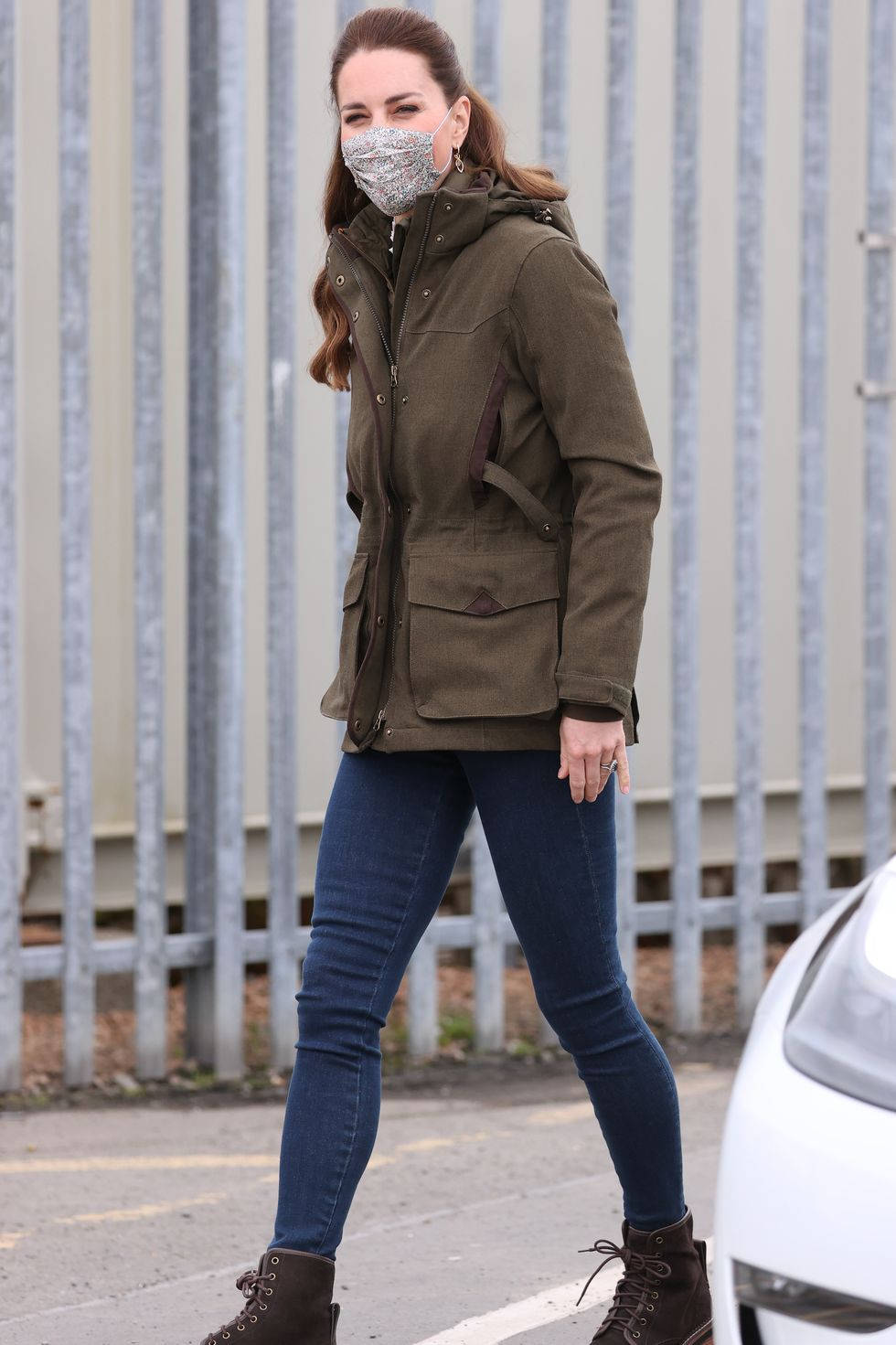 Kate Middleton 桌布 壁紙 — 一位身穿橄欖色外套和牛仔褲的女性在戶外靠近金屬圍欄的地方行走，一輛白色汽車部分可見。