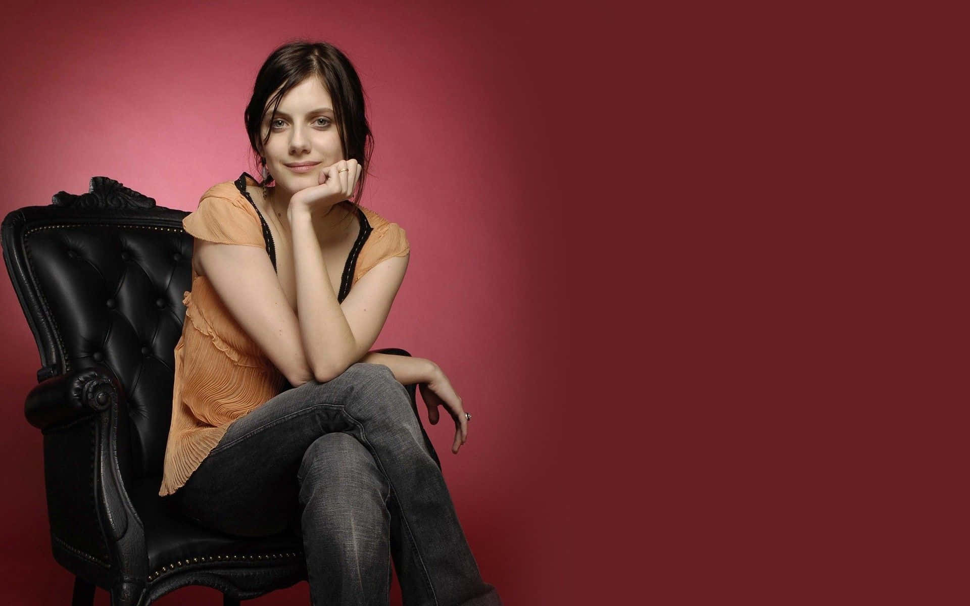 Mélanie Laurent 桌布 壁紙 — 一位身穿橙色上衣和牛仔褲的女性，放鬆地坐在黑色皮椅上，背景是深紅色的攝影棚佈景。