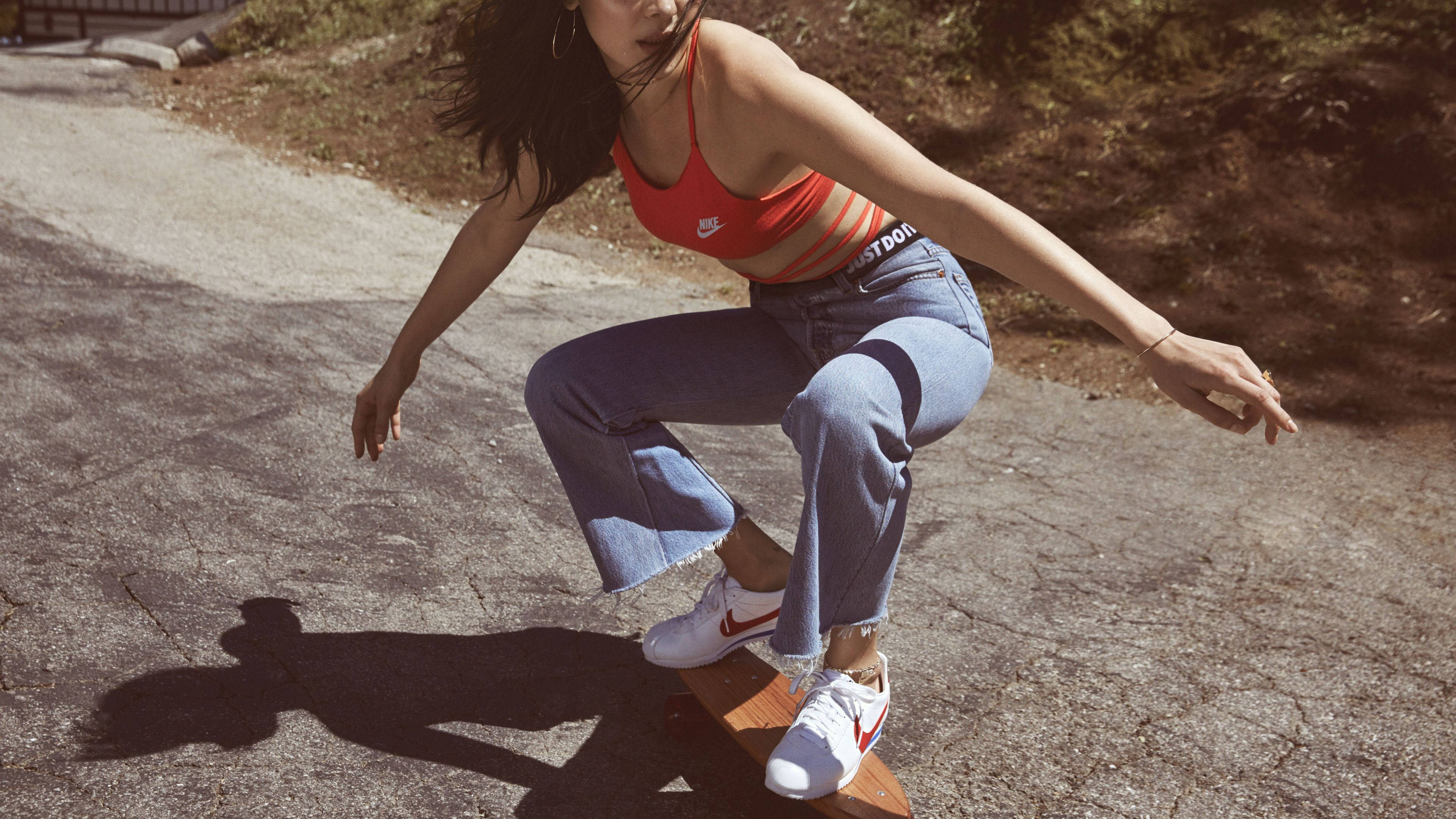 nike girl 桌布 壁紙 — 一位身穿橙色上衣和藍色牛仔褲的年輕女子低蹲在滑板上，在明亮的陽光下在裂開的路面上投下清晰的影子。