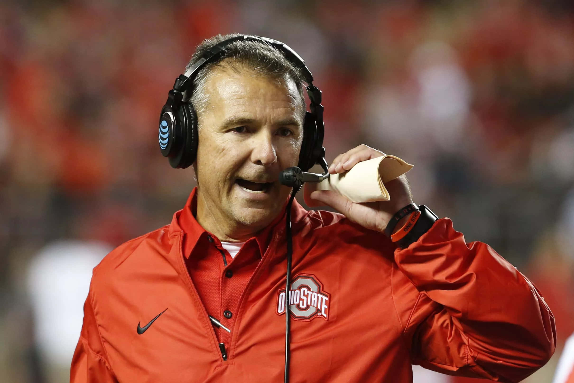 Urban Meyer 桌布 壁紙 — 一位身穿橙色俄亥俄州立大學外套的美式足球教練在比賽中對著耳機麥克風大喊，身後是模糊的體育場人群。
