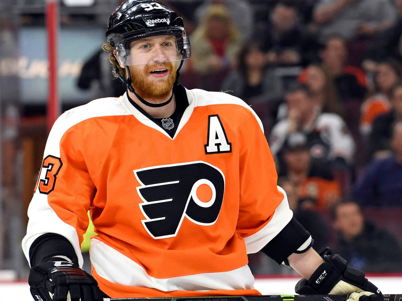 hockey 桌布 壁紙 — 一位身穿橙色和白色球衣的費城飛人隊冰球球員在比賽中微笑，背景是模糊的球迷。