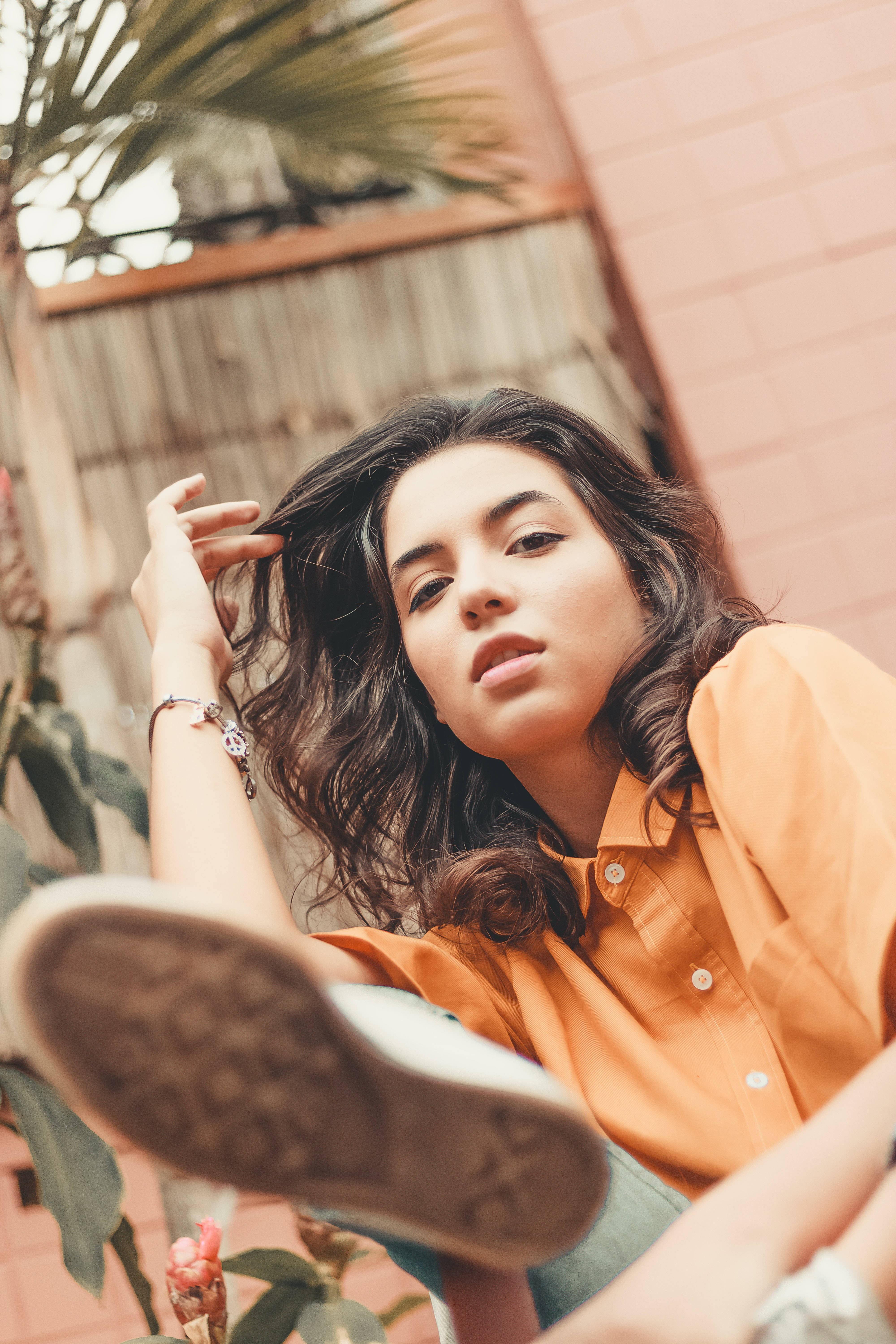 Latina Girl 桌布 壁紙 — 一位身穿橙色襯衫、深色波浪髮的年輕女子，隨意地靠在一面有熱帶植物的粉紅色磚牆上。