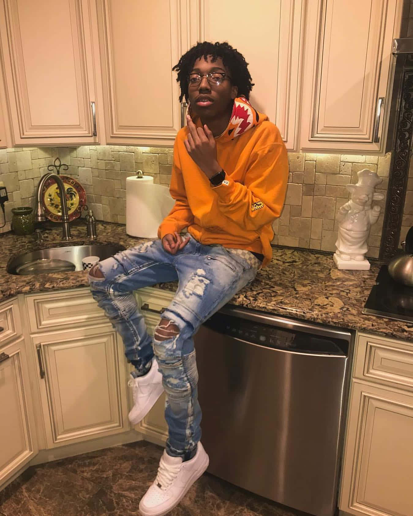 Rappers 桌布 壁紙 — 一位身穿橙色連帽衫和破洞牛仔褲的年輕男子隨意地坐在廚房檯面上，下方是不鏽鋼洗碗機。