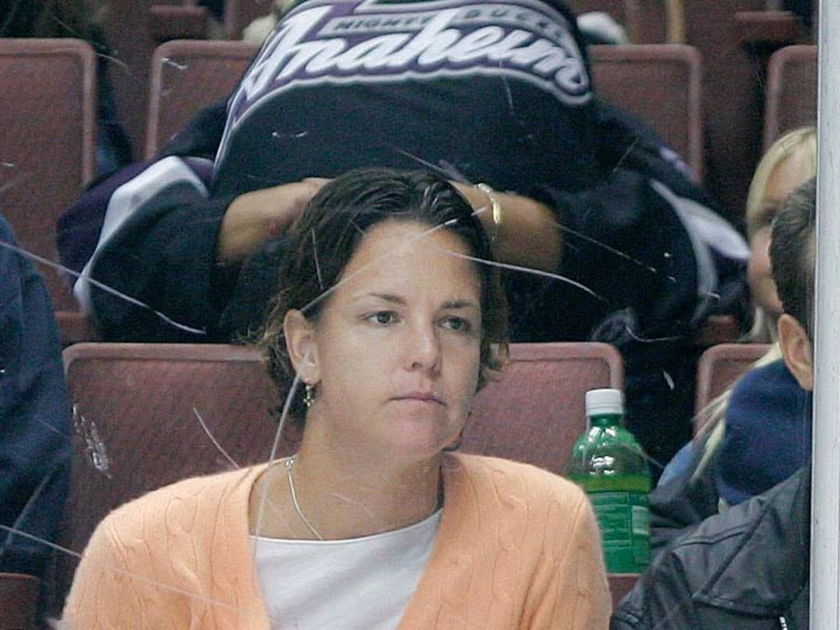 Tennis 桌布 壁紙 — 一位身穿橙色開襟毛衣的女性坐在體育場座位後方，隔著一道破裂的玻璃屏障，附近有一個綠色瓶子。