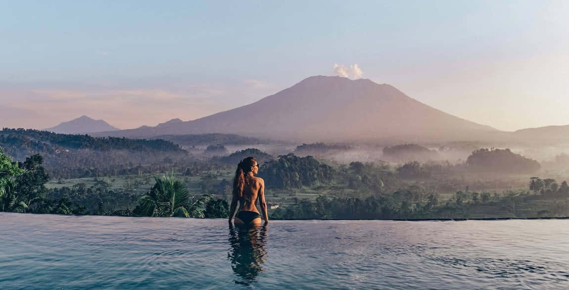 Bali Island 桌布 壁紙 — 一位身穿比基尼的女士站在無邊際泳池中，欣賞日出時霧氣繚繞的山脈。