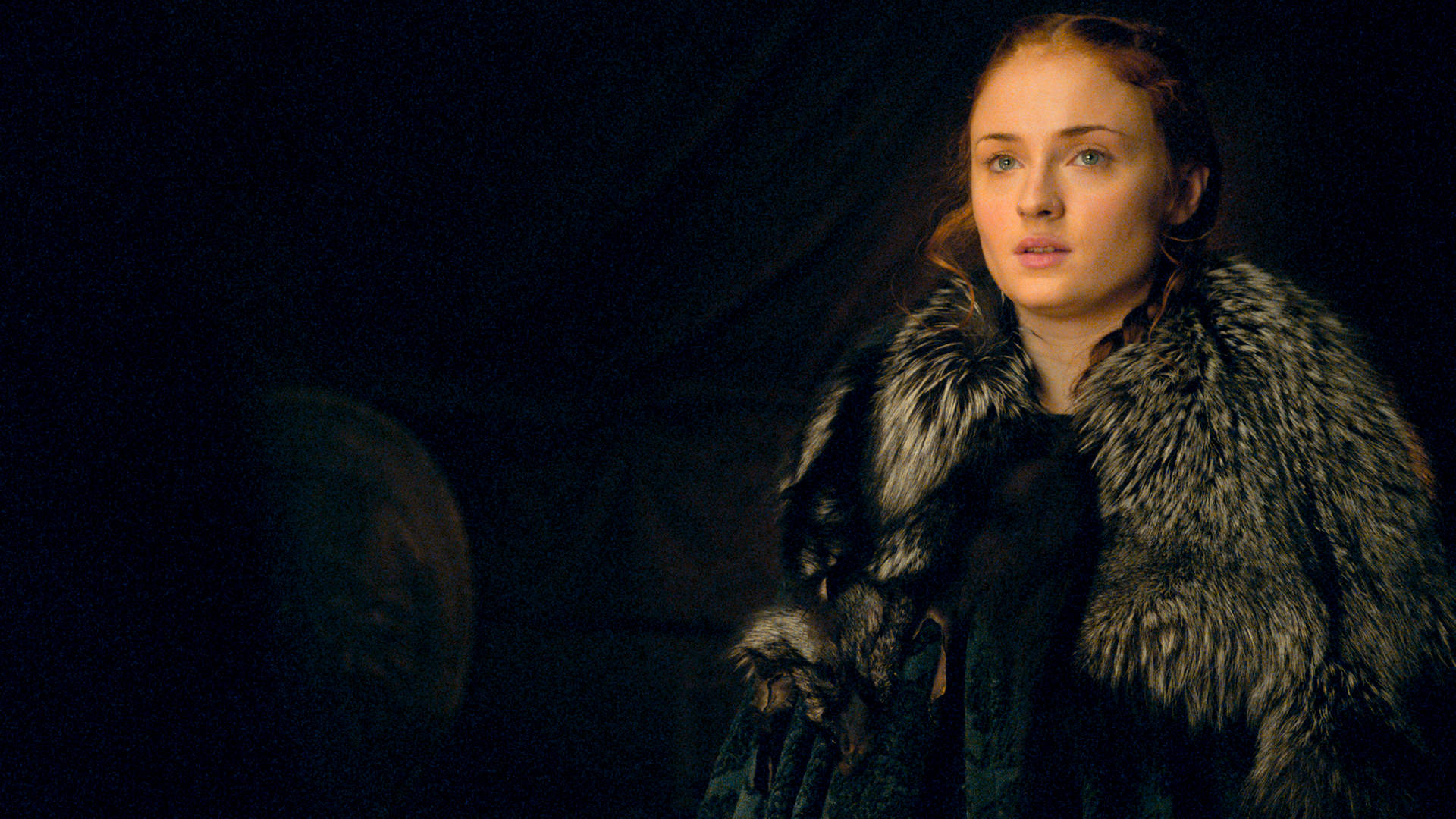Sansa Stark 桌布 壁紙 — 一位身穿毛皮襯裡斗篷的女子在昏暗光線下，對著黑暗背景專注地凝視。