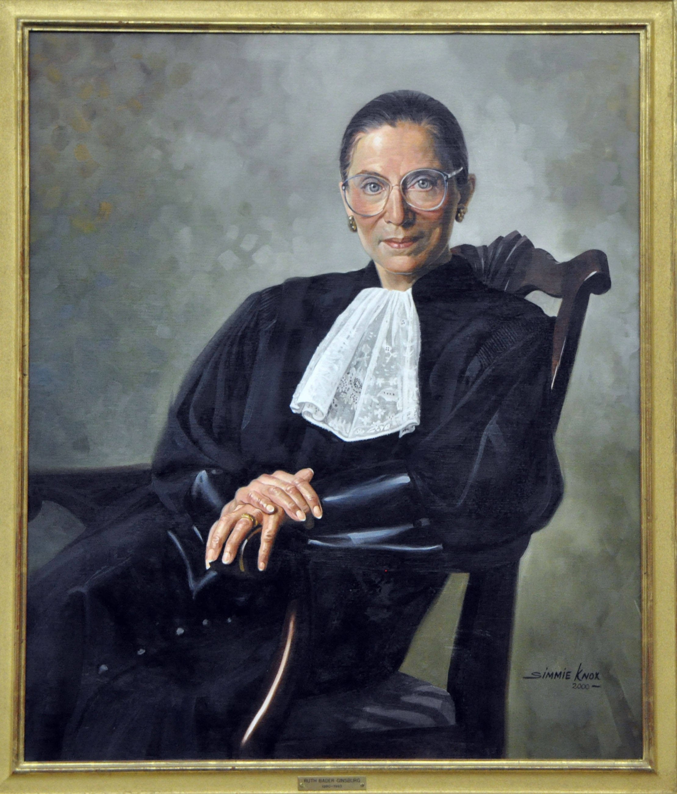 Ruth Bader Ginsburg 桌布 壁紙 — 一位身穿法官袍、戴眼鏡的女性坐在木椅上，背景為柔和灰色的正式肖像。