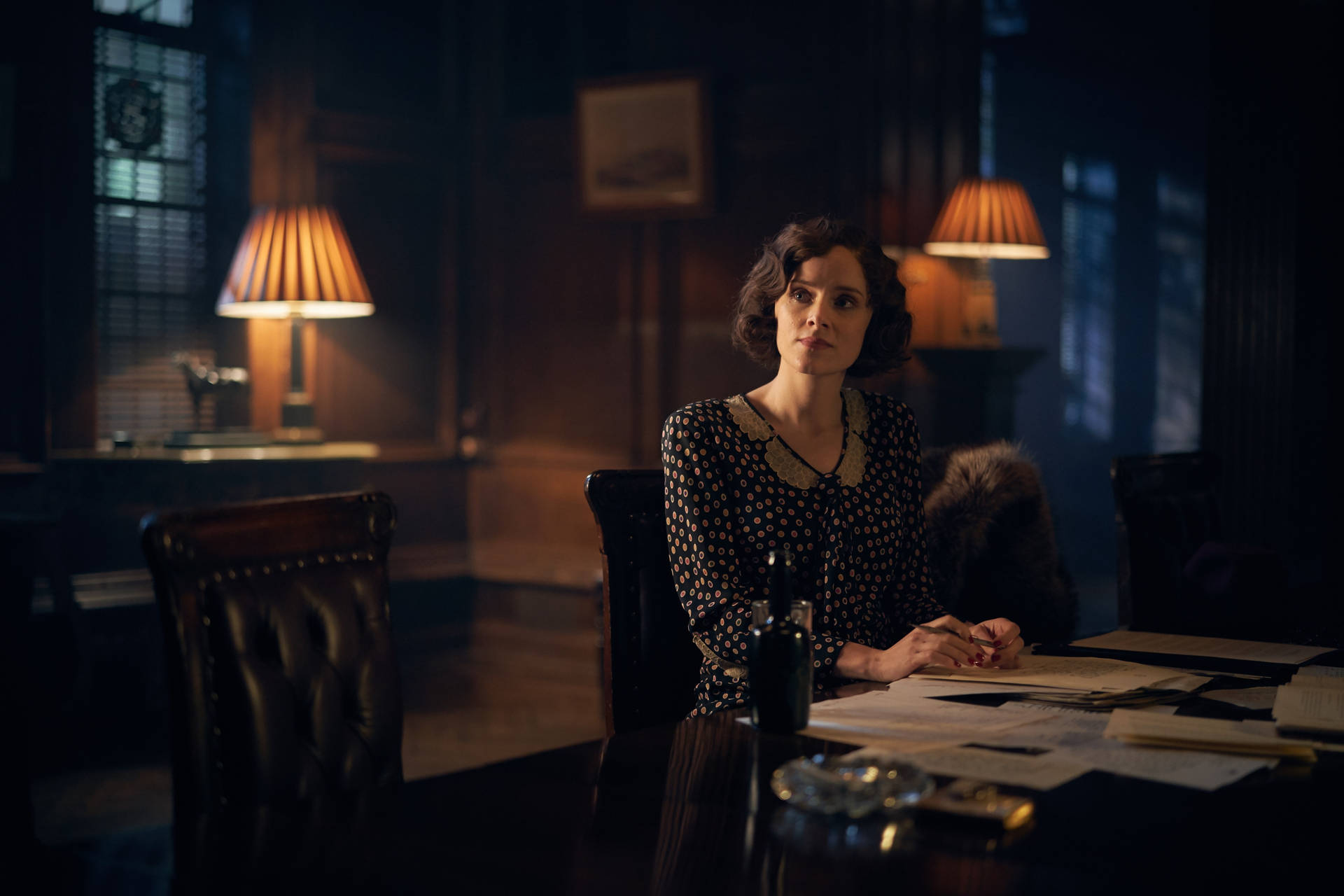 身穿波點連衣裙的女性坐在昏暗復古辦公室的木桌旁。 — Peaky Blinders 壁紙