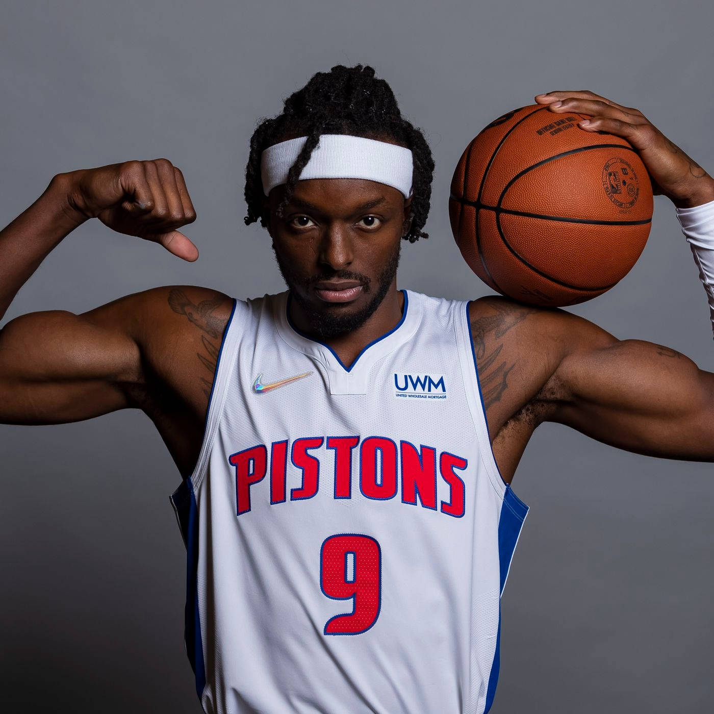 NBA 桌布 壁紙 — 一位身穿活塞隊球衣的肌肉發達籃球運動員，在灰色工作室背景前自信地展示肌肉，同時手持籃球。