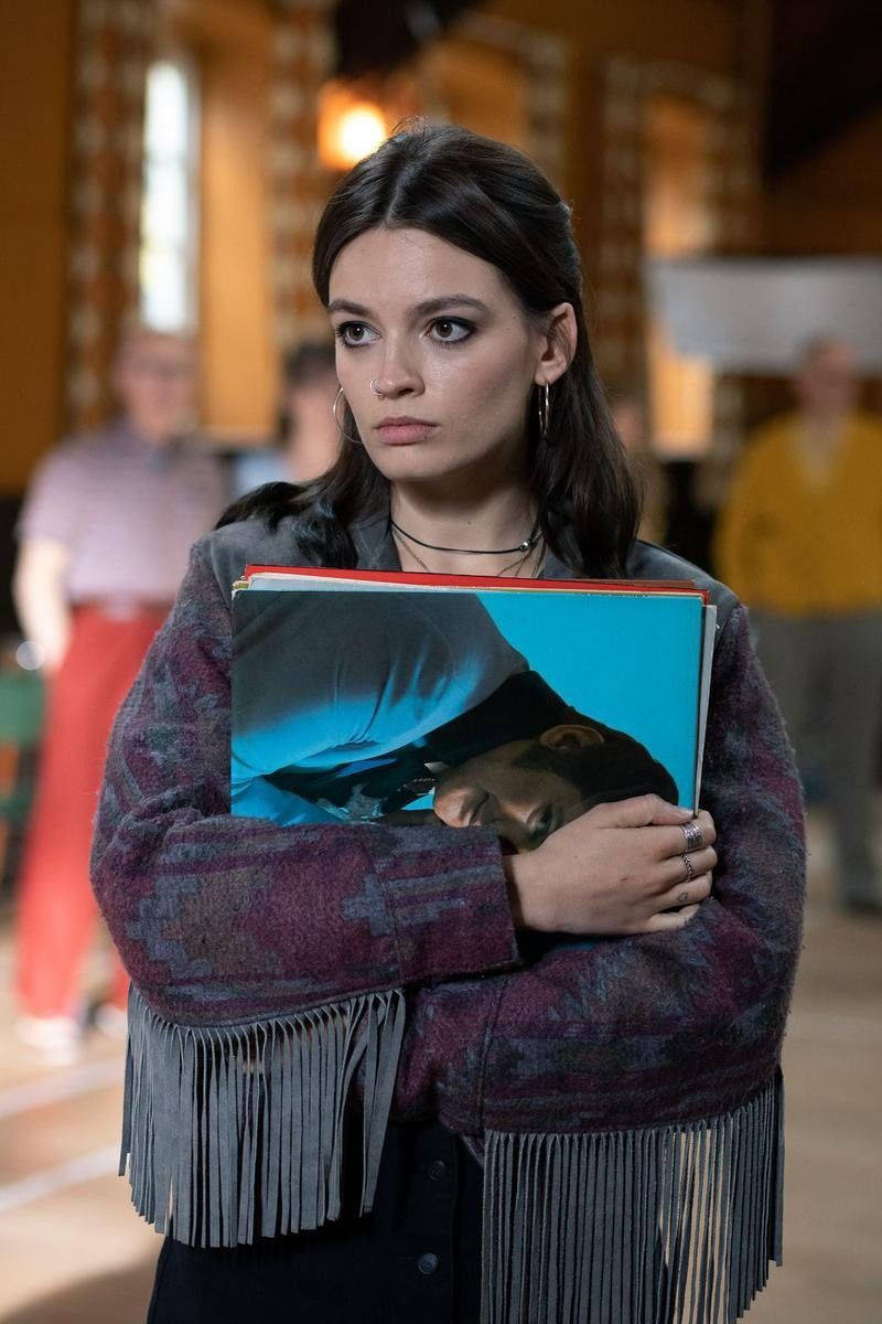 maeve wiley 桌布 壁紙 — 一位身穿流蘇外套的年輕女性，在溫暖照明的室內空間中，身後有模糊的人群，手持黑膠唱片專輯。
