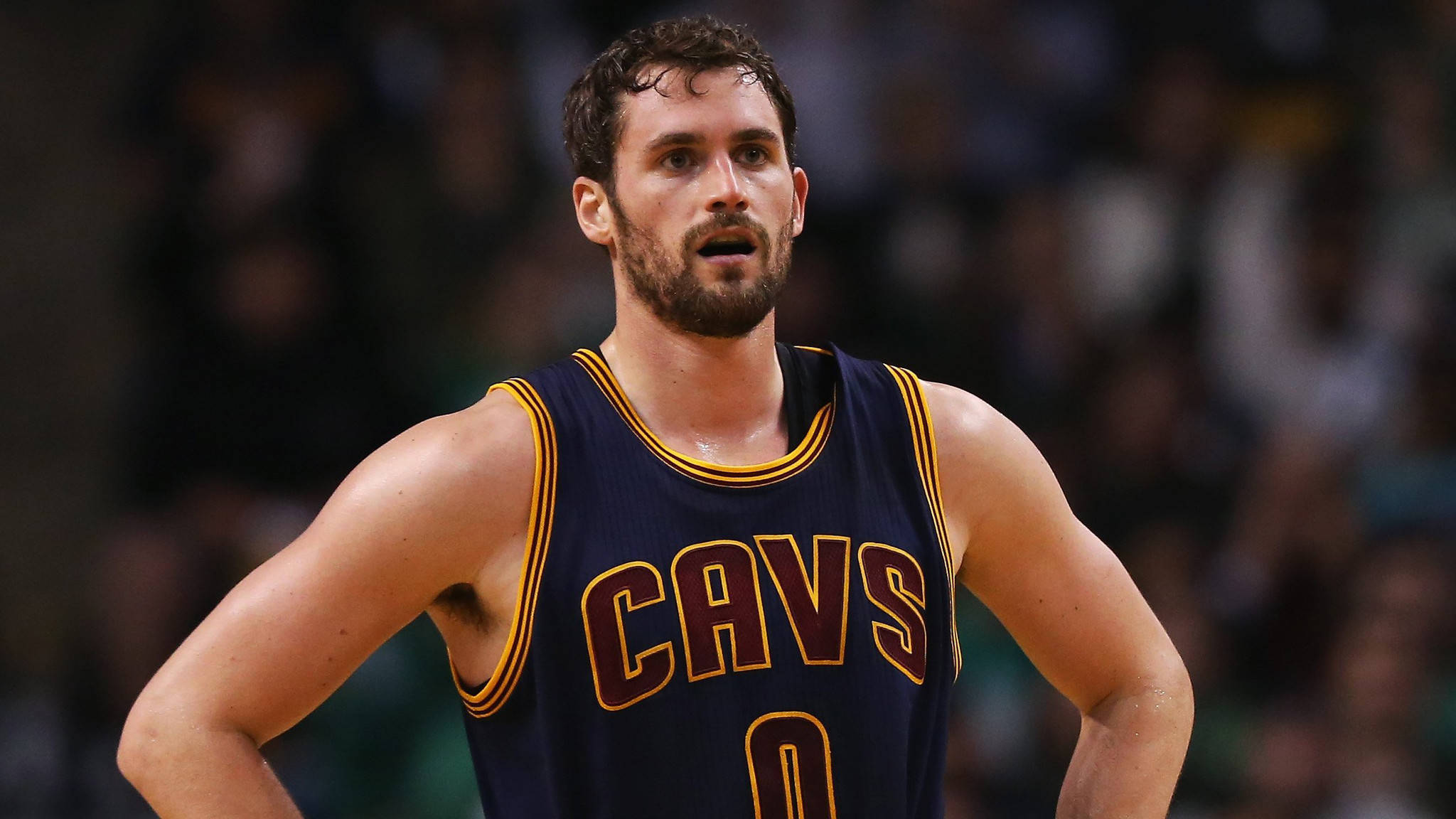 Kevin Love 桌布 壁紙 — 一位身穿海軍藍Cavs球衣的專注籃球運動員，在明亮的場館燈光下比賽中雙手叉腰站立。