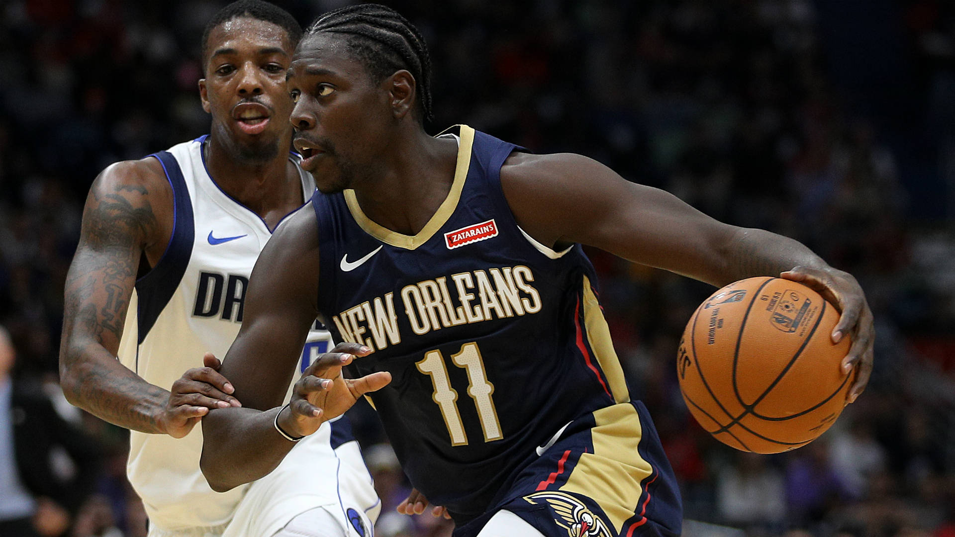 Jrue Holiday 桌布 壁紙 — 一位身穿海軍藍和金色球衣的紐奧良鵜鶘球員在激烈的NBA比賽中運球，同時被一位身穿白色球衣的達拉斯獨行俠球員緊密防守。