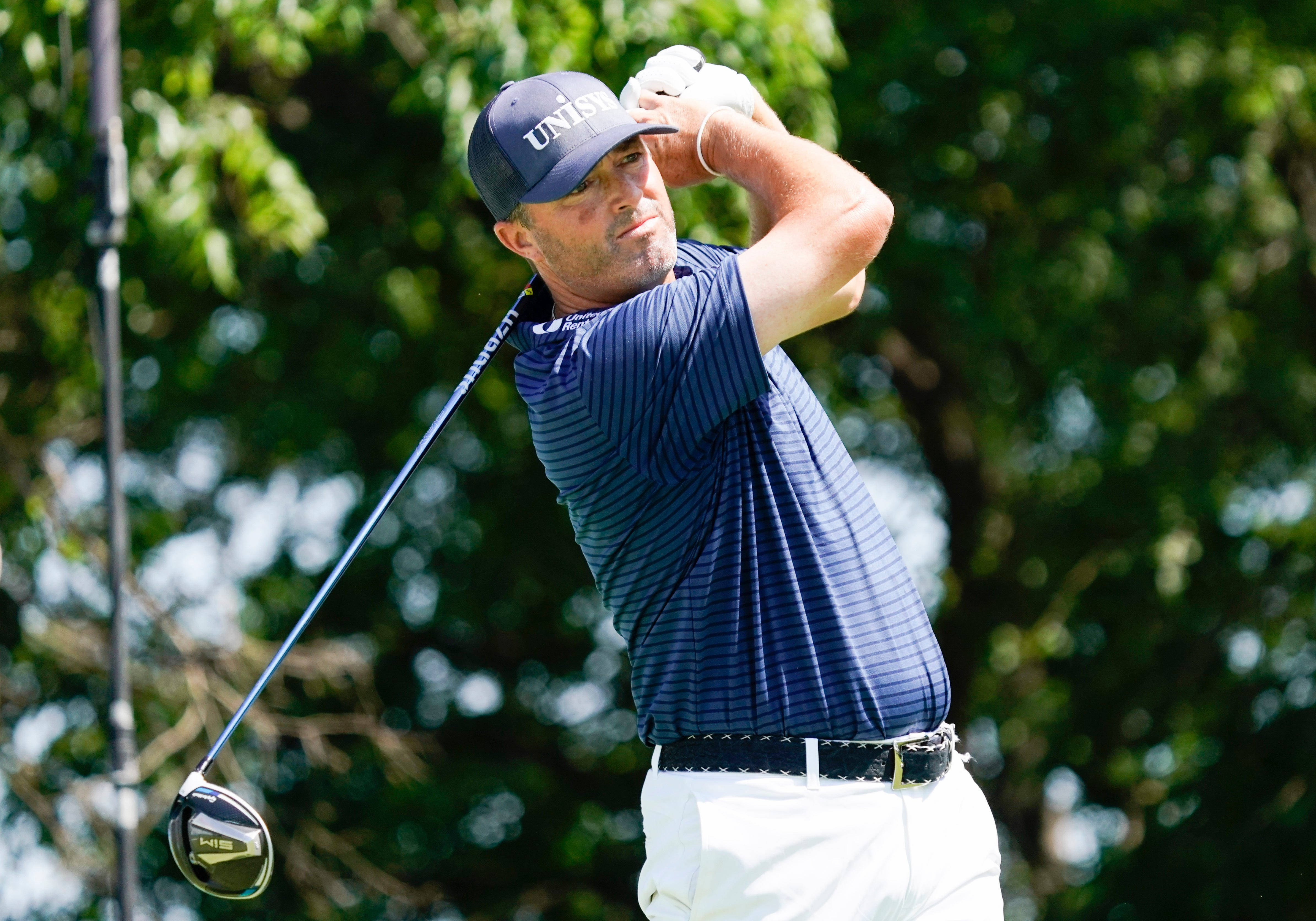 Ryan Palmer 桌布 壁紙 — 一位身穿海軍藍條紋襯衫和白色褲子的男性高爾夫球手，在明亮的陽光下，以茂密的綠樹為背景，揮出強力的一桿。