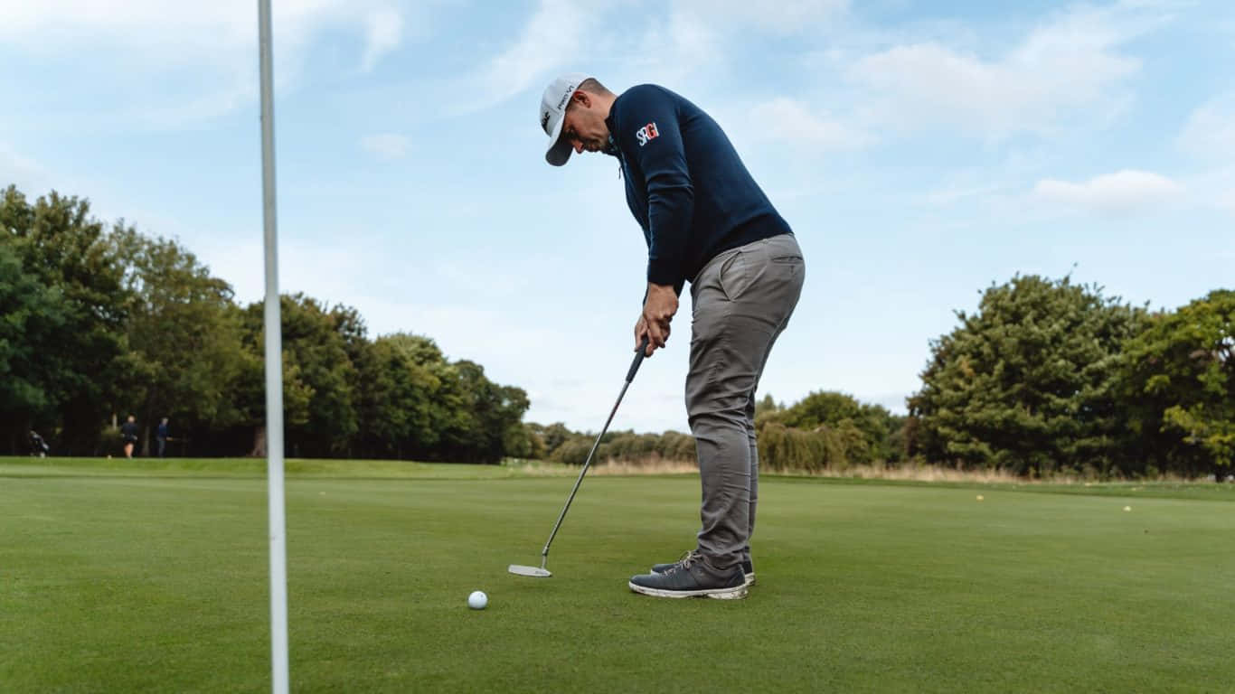 1366x768 Golf Course 桌布 壁紙 — 一位身穿海軍藍毛衣的高爾夫球手在晴朗藍天下，專注地將白色球推入茂密綠色球道上的洞口附近。