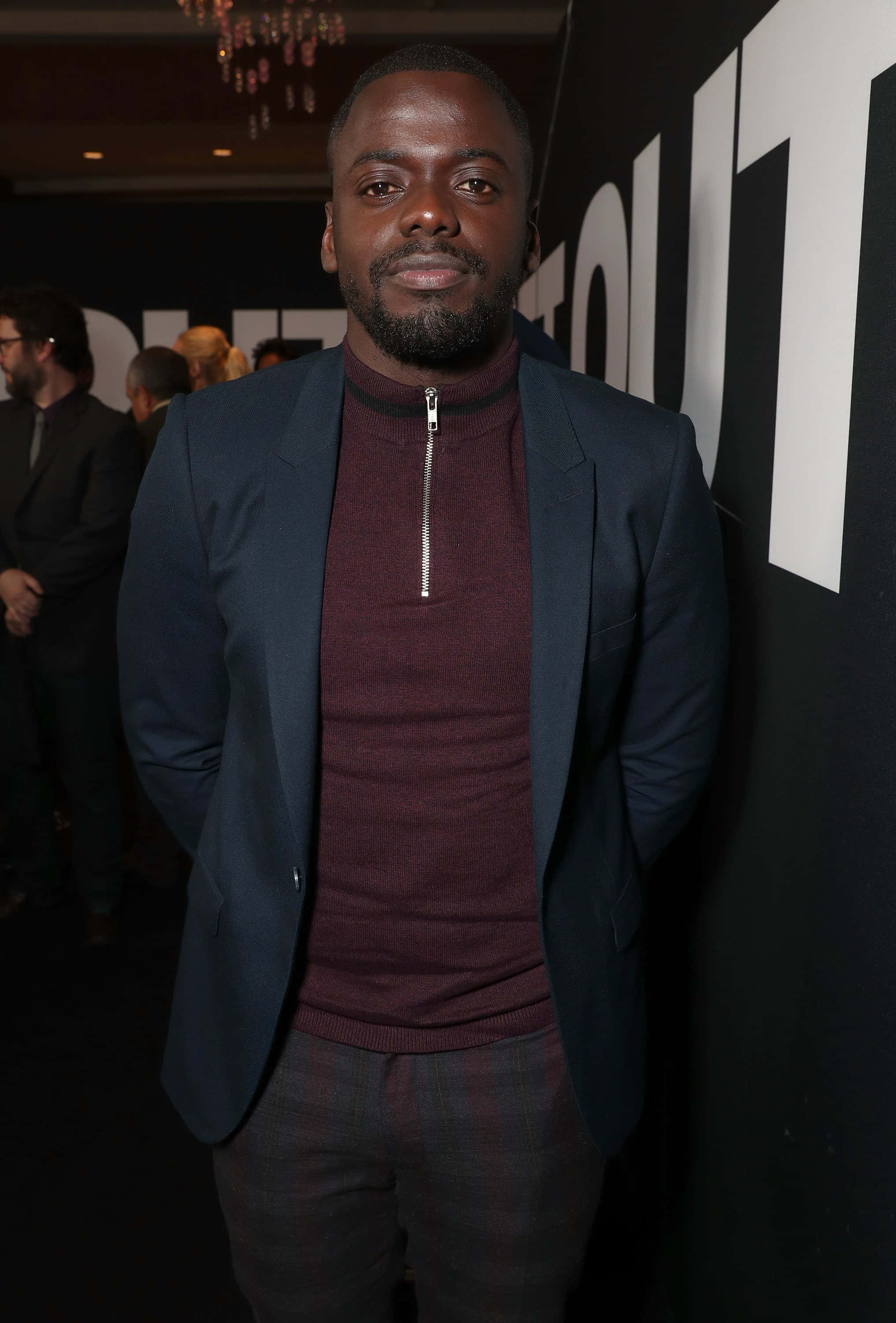 Daniel Kaluuya 桌布 壁紙 — 一位身穿海軍藍西裝外套和栗色拉鍊高領毛衣的男子，在一個有吊燈的活動中自信地站立。