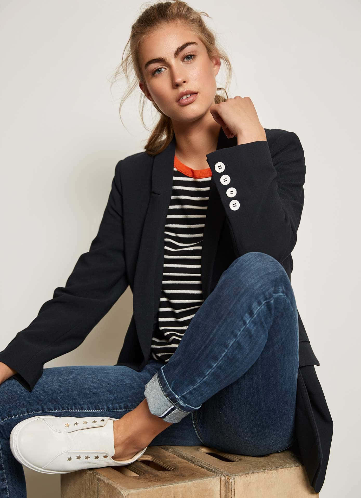 Navy Blue Blazer 桌布 壁紙 — 一位身穿海軍藍西裝外套和條紋T恤的年輕女性，隨意地坐在白色背景前的木箱上。