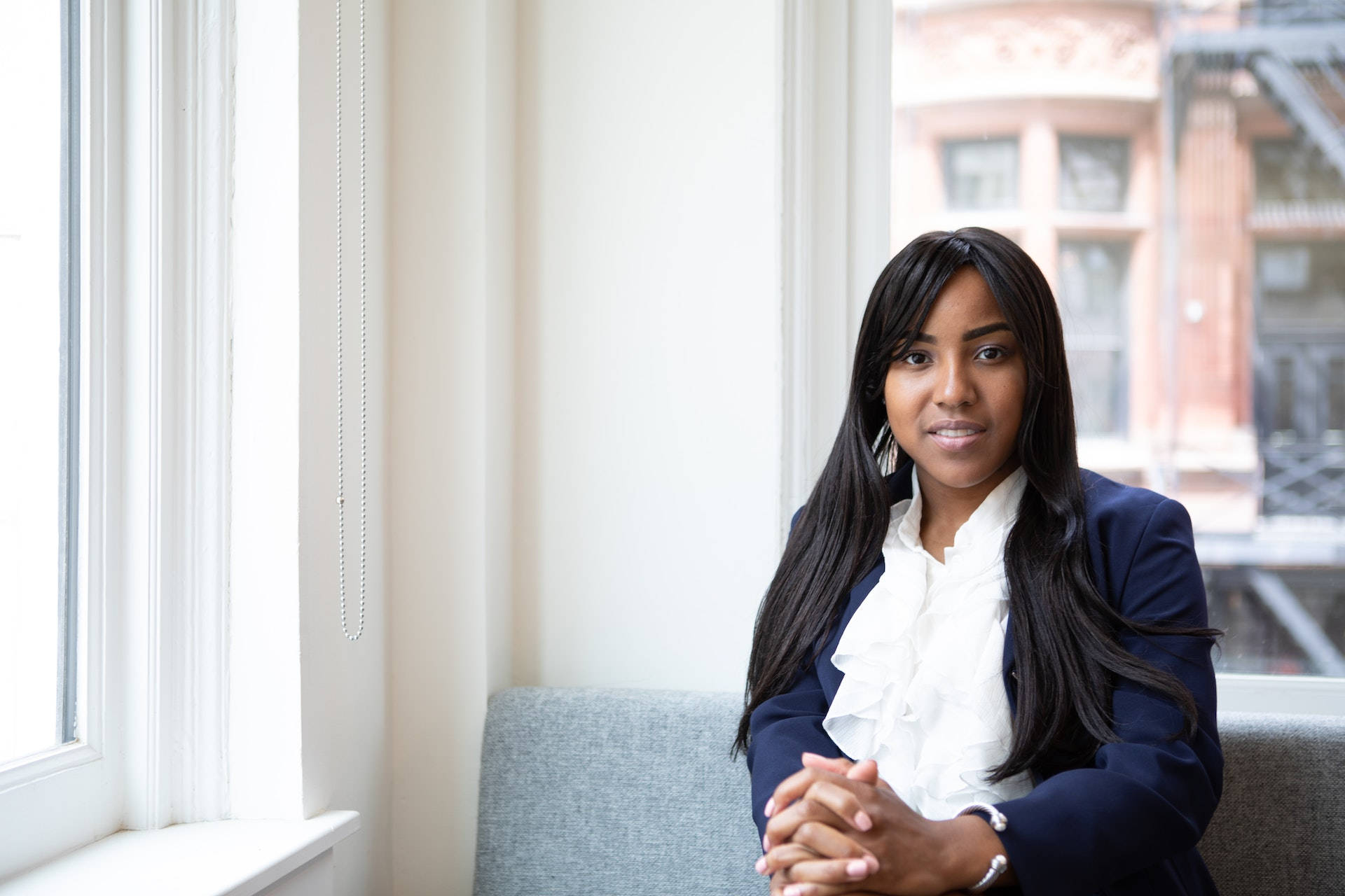 Black Girl 桌布 壁紙 — 一位身穿海軍藍西裝外套和白色襯衫的沉著女性坐在明亮的窗邊，雙手緊握，直視鏡頭。