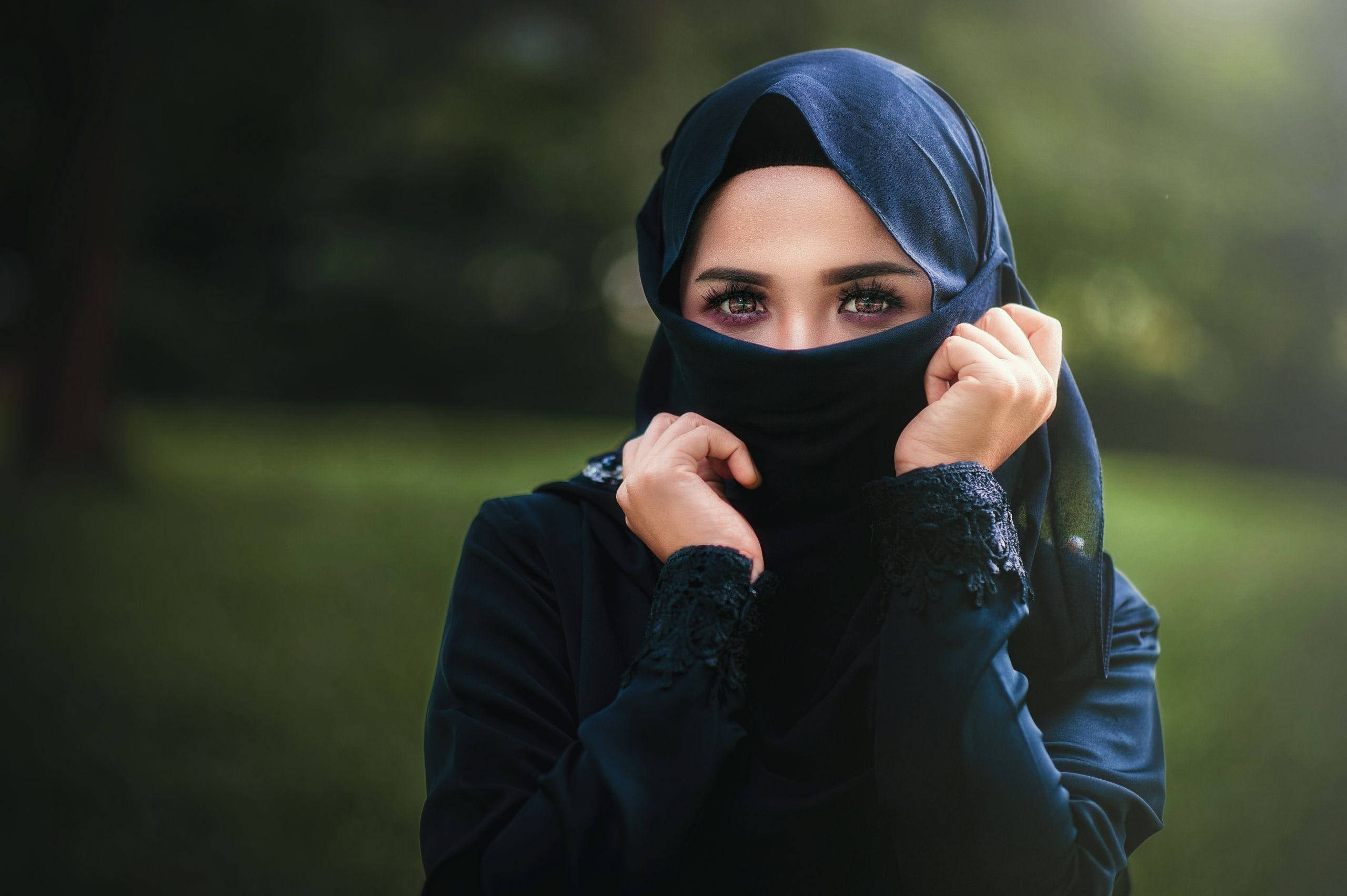muslim 桌布 壁紙 — 一位身穿海軍藍頭巾的女性輕輕掀起面紗，在柔和的綠色公園背景下露出富有表現力的眼睛。