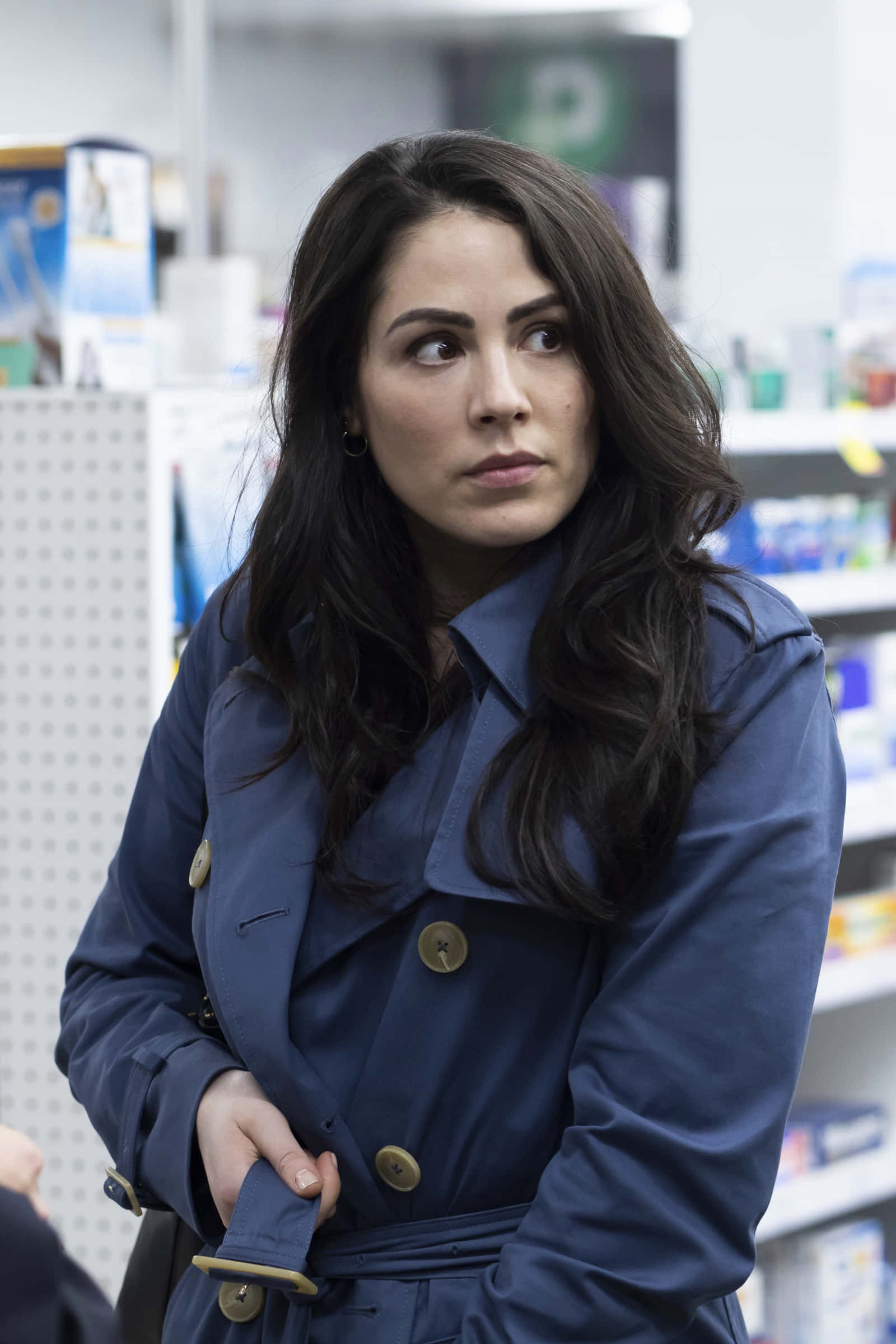 Michelle Borth 桌布 壁紙 — 一位身穿海軍藍風衣的女性站在藥局走道上，側目露出擔憂的表情。