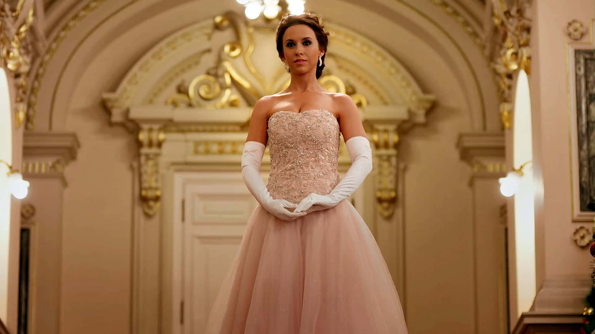 lacey chabert 桌布 壁紙 — 一位身穿淡粉紅色舞會禮服和白色手套的女性，優雅地站在一個帶有金色點綴和溫暖燈光的華麗宏偉走廊中。