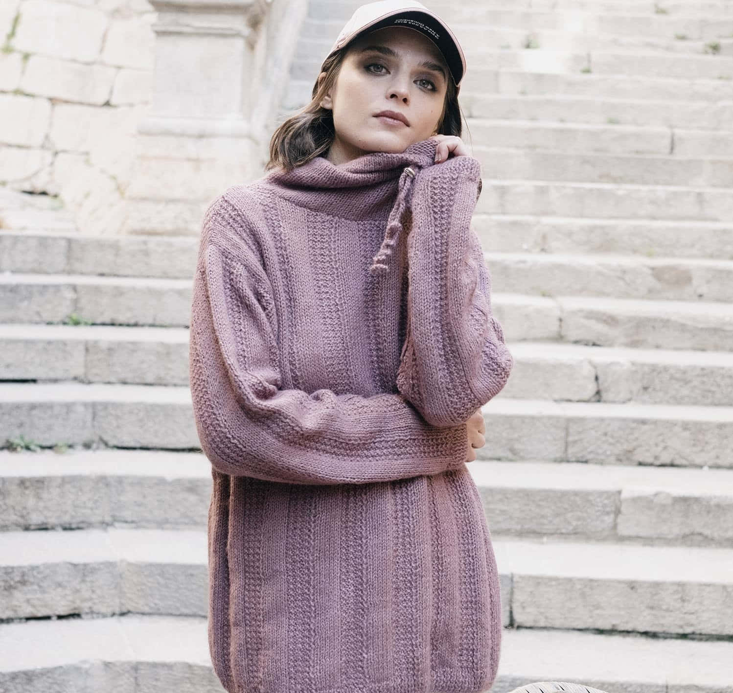 Purple Turtle-neck Sweater 桌布 壁紙 — 一位身穿淡紫色針織毛衣和帽子的年輕女子，在柔和日光下若有所思地站在古老的石階上。