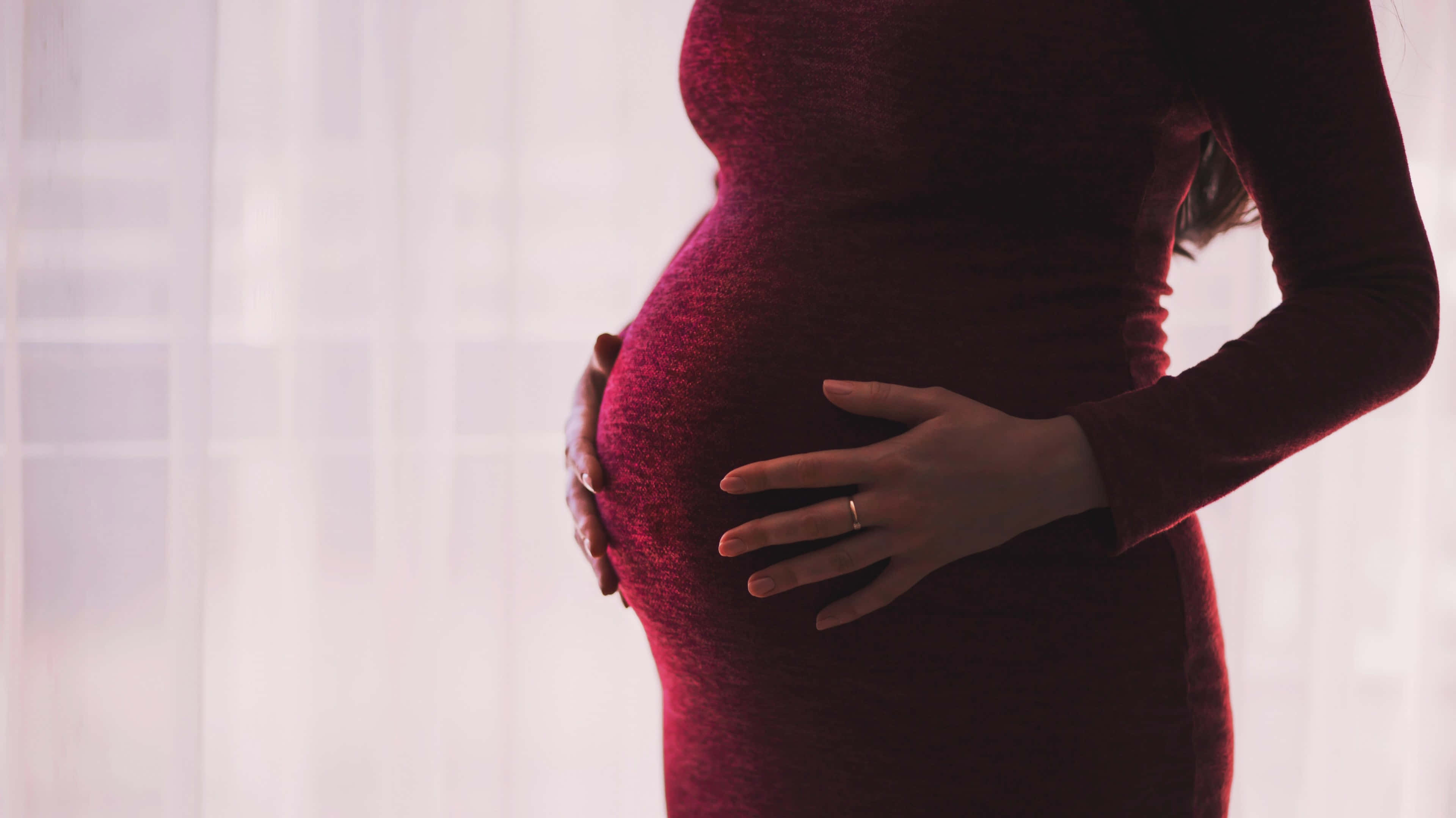 Pregnant 桌布 壁紙 — 一位身穿深紅色連衣裙的孕婦，在柔軟的白色窗簾背景下輕輕地捧著她的肚子。