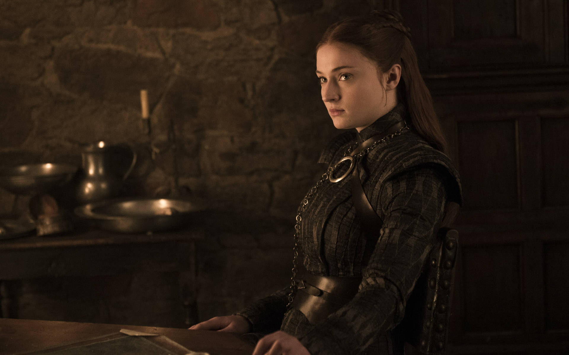 Sansa Stark 桌布 壁紙 — 一位身穿深色中世紀盔甲的女性坐在一張木桌旁，房間內有石牆和燭光，光線昏暗。