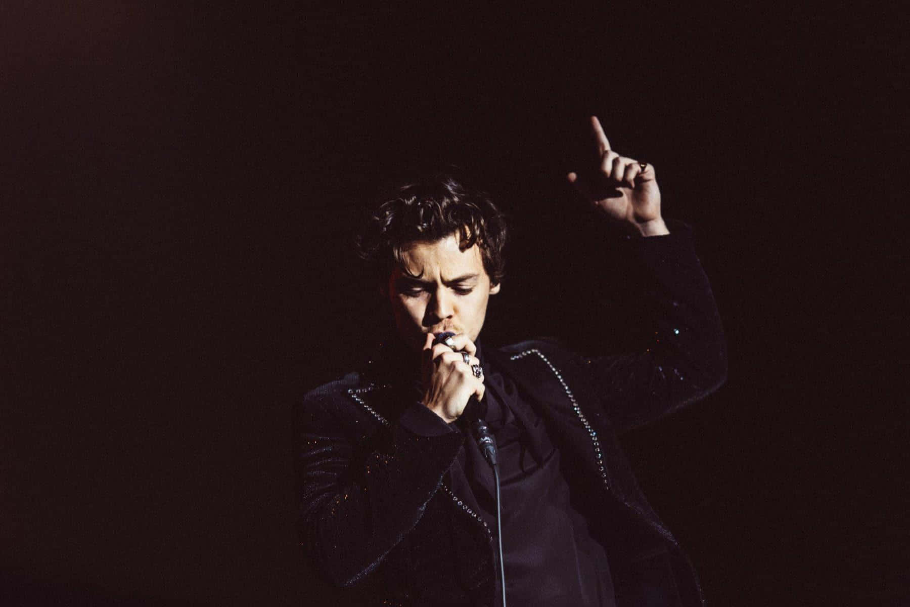 Harry Styles Laptop 桌布 壁紙 — 一位身穿深色亮片外套的男歌手在戲劇性的舞台燈光下熱情地對著麥克風演唱。