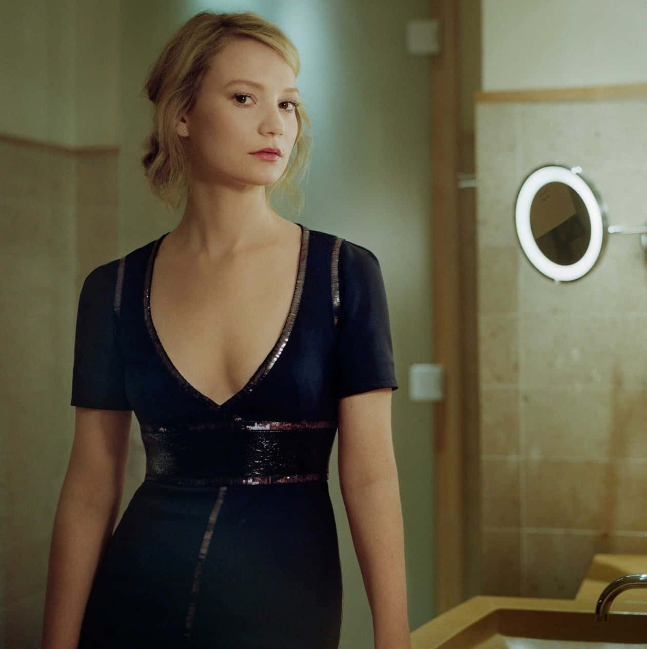 Mia Wasikowska 桌布 壁紙 — 一位身穿深色亮片連衣裙的女性站在一個光線柔和、配有圓鏡和木製裝置的浴室中。