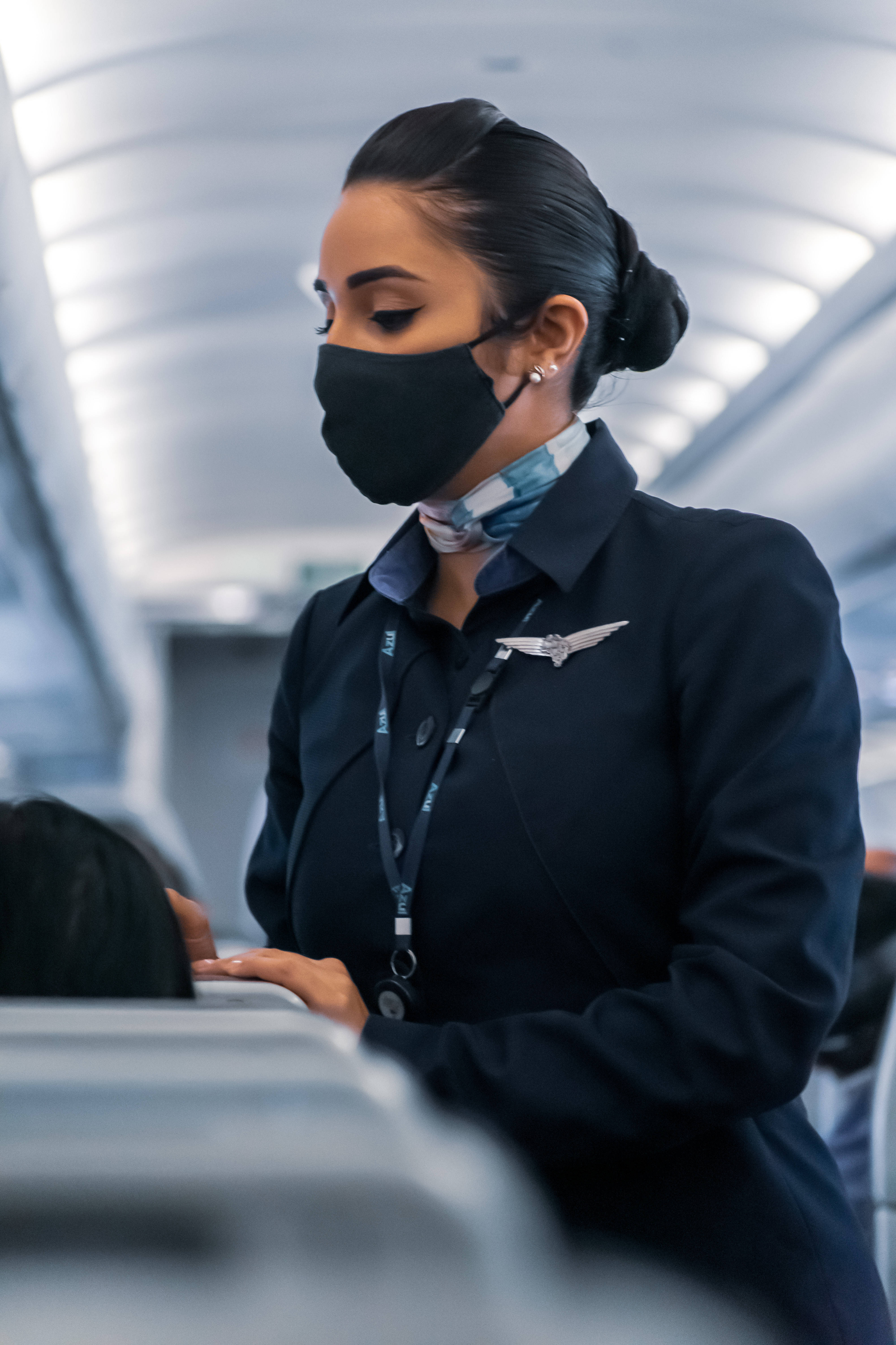 Airport 桌布 壁紙 — 一位身穿深色制服和黑色口罩的空中服務員正在現代飛機艙內為乘客服務。