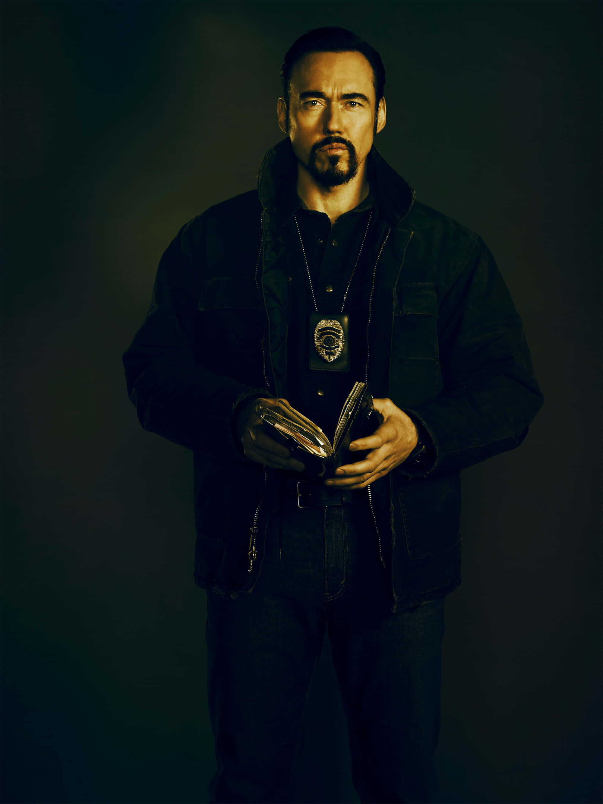 Kevin Durand 桌布 壁紙 — 一位身穿深色外套的嚴肅警察，在陰暗陰影的背景前拿著徽章和錢包。