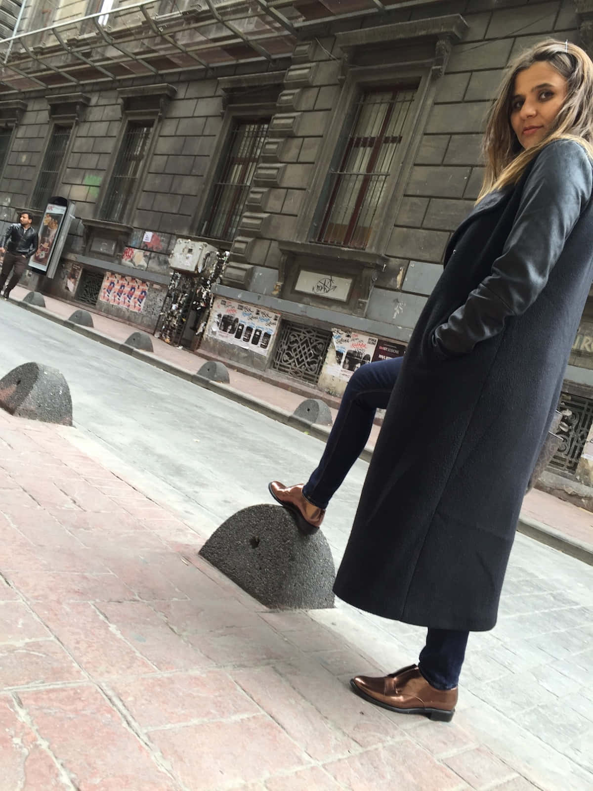 鞋子 桌布 壁紙 — 一位身穿深色外套的女性在歷史建築附近的磚砌城市人行道上，在石柱上保持平衡。