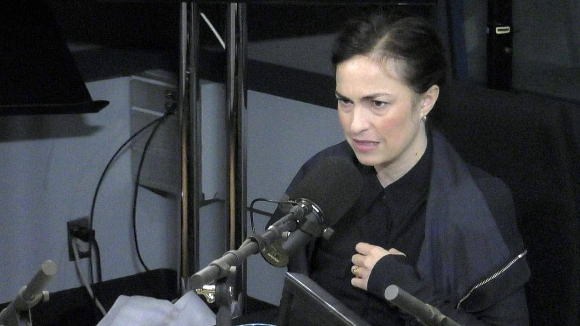 Woman 桌布 壁紙 — 一位身穿深色外套的女性坐在一張有麥克風的桌子前，身處燈光昏暗的錄音室環境中。