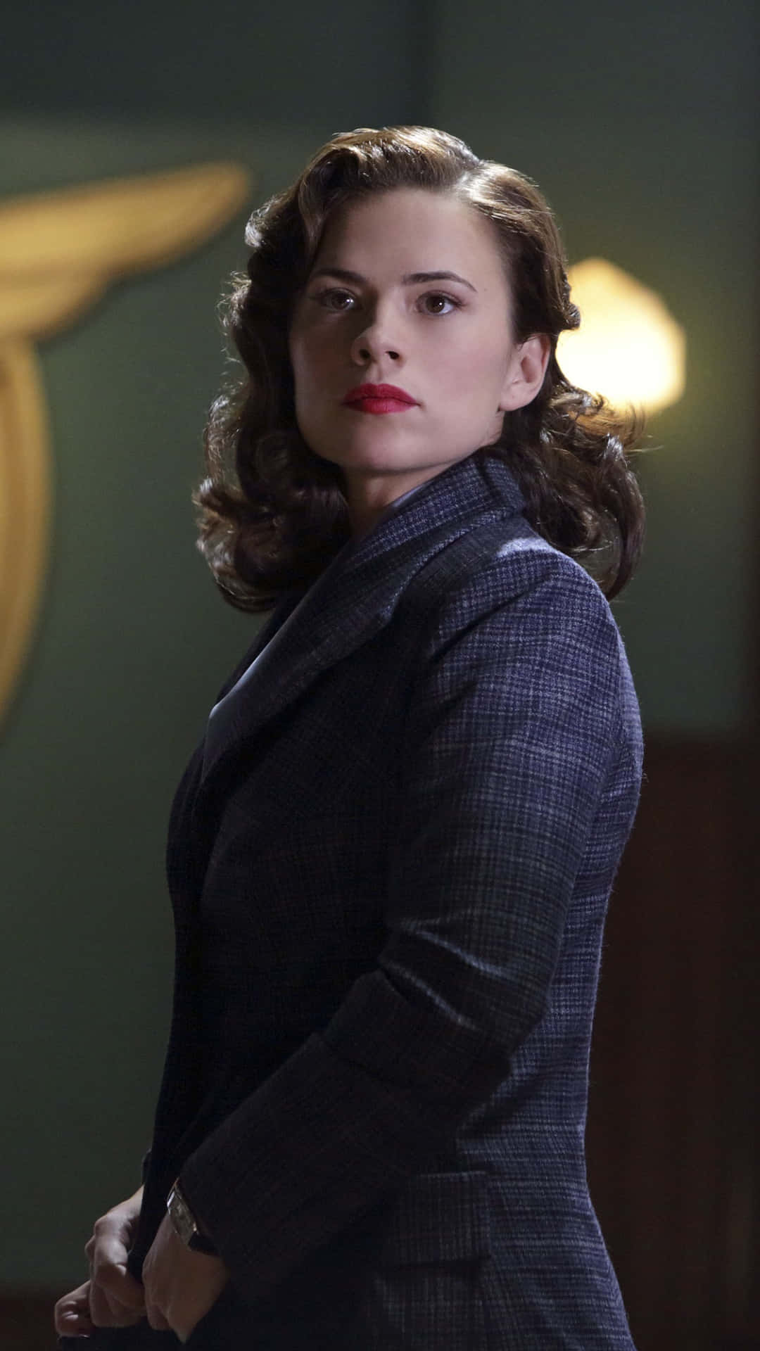 Peggy Carter 桌布 壁紙 — 一位身穿深色格紋西裝外套的女性，站在燈光昏暗的房間裡，身後有一盞溫暖的燈和一個金色帶翼的徽章。