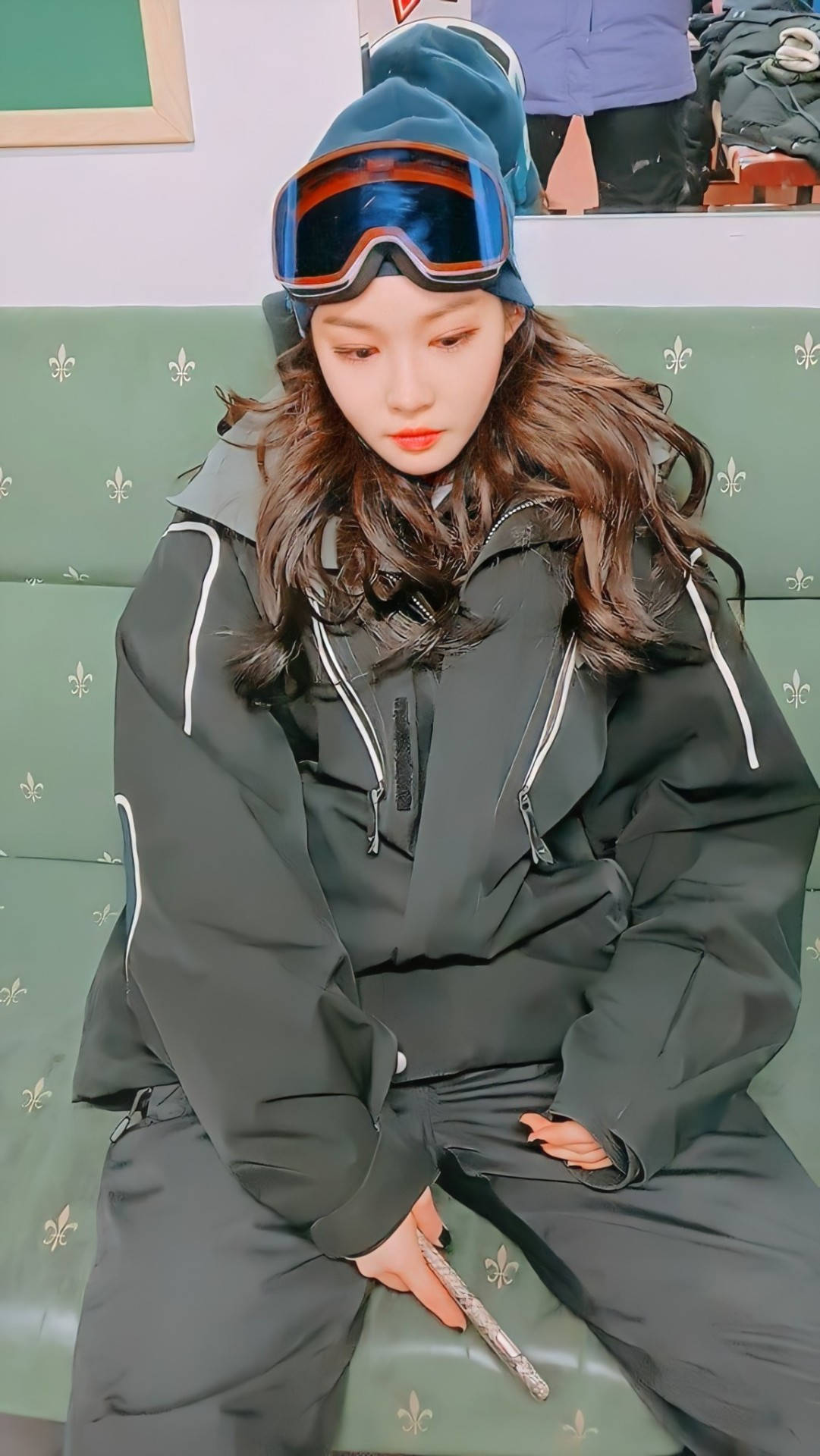 Chungha 桌布 壁紙 — 一位身穿深色滑雪服和護目鏡的年輕女性，若有所思地坐在一張綠色鳶尾花圖案的沙發上。