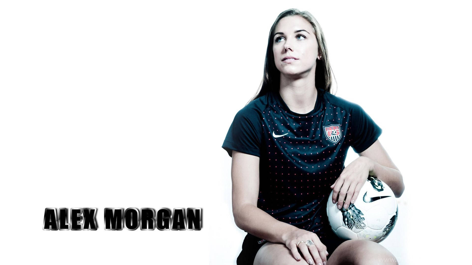 Alex Morgan 桌布 壁紙 — 一位身穿深色球衣的女足球員拿著球，仰望純白背景。