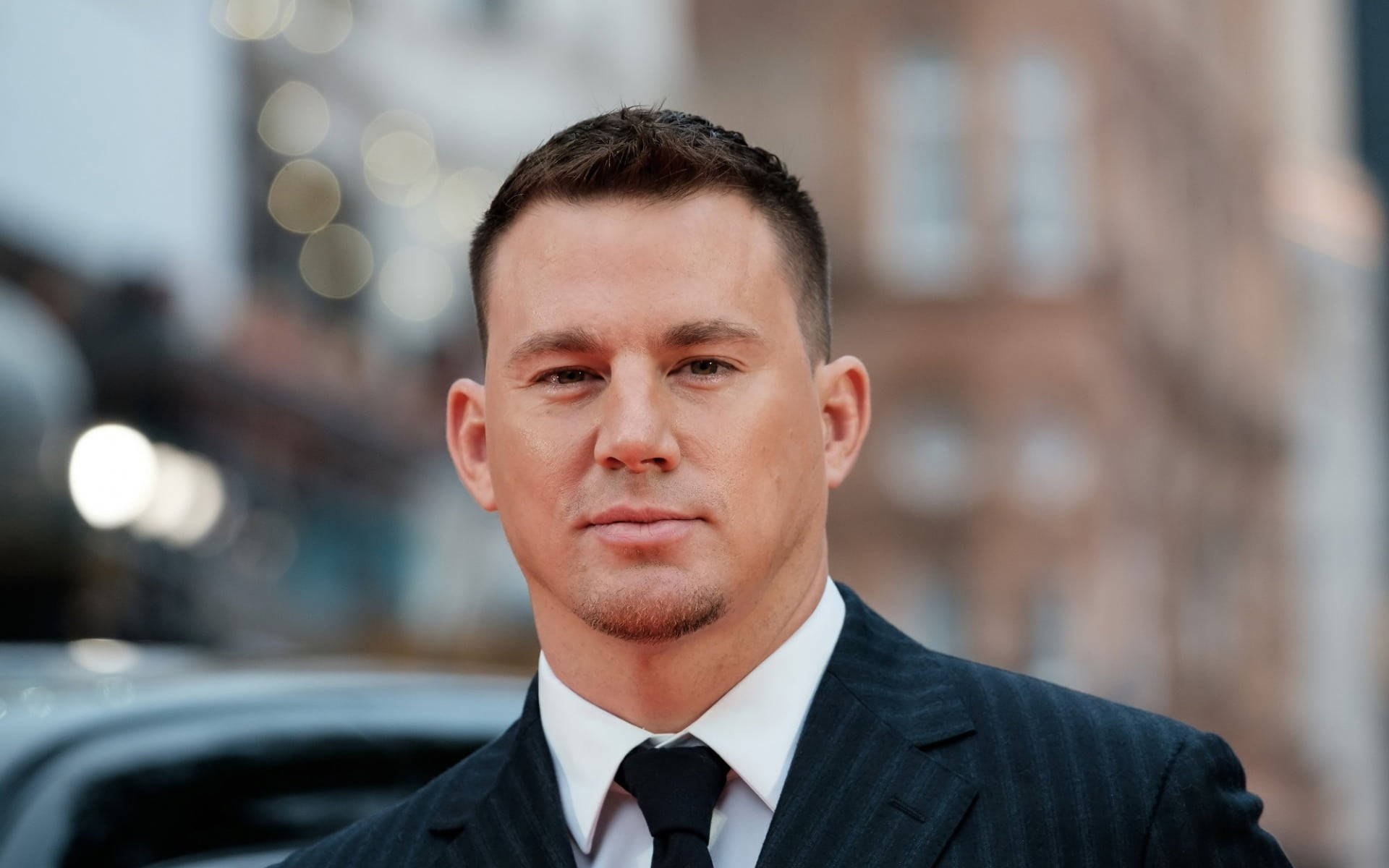 Channing 桌布 壁紙 — 一位身穿深色細條紋西裝和黑色領帶的男士的特寫肖像，戶外站立，背景是模糊的城市燈光和建築物。