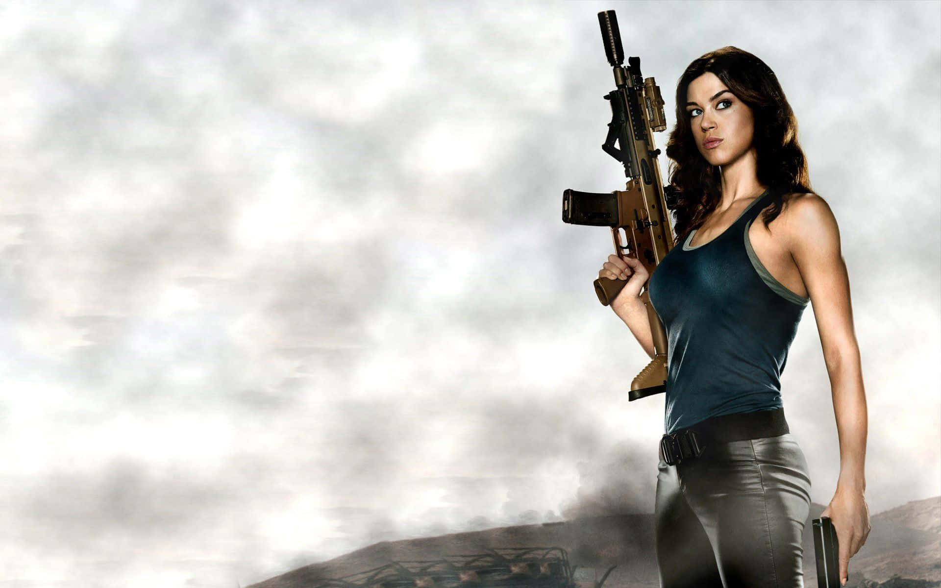 Adrianne Palicki 桌布 壁紙 — 一位身穿深色背心和褲子的女性，在陰天和遠山背景下持步槍。