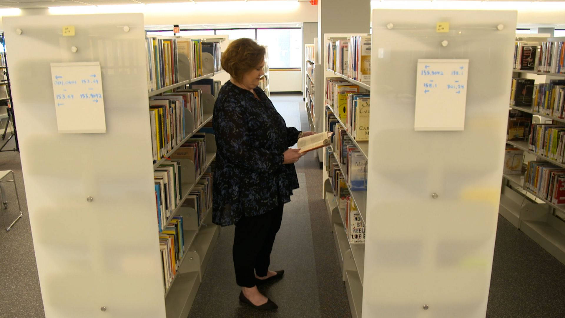 Library 桌布 壁紙 — 一位身穿深色花卉襯衫的女性在安靜的圖書館通道中瀏覽書籍，通道兩旁是高高的白色書架。
