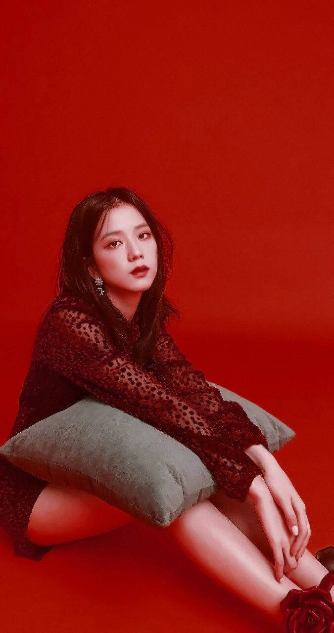 Jisoo Cute 桌布 壁紙 — 一位身穿深色蕾絲上衣的女性，優雅地坐在綠色枕頭上，背景是鮮明的紅色攝影棚。