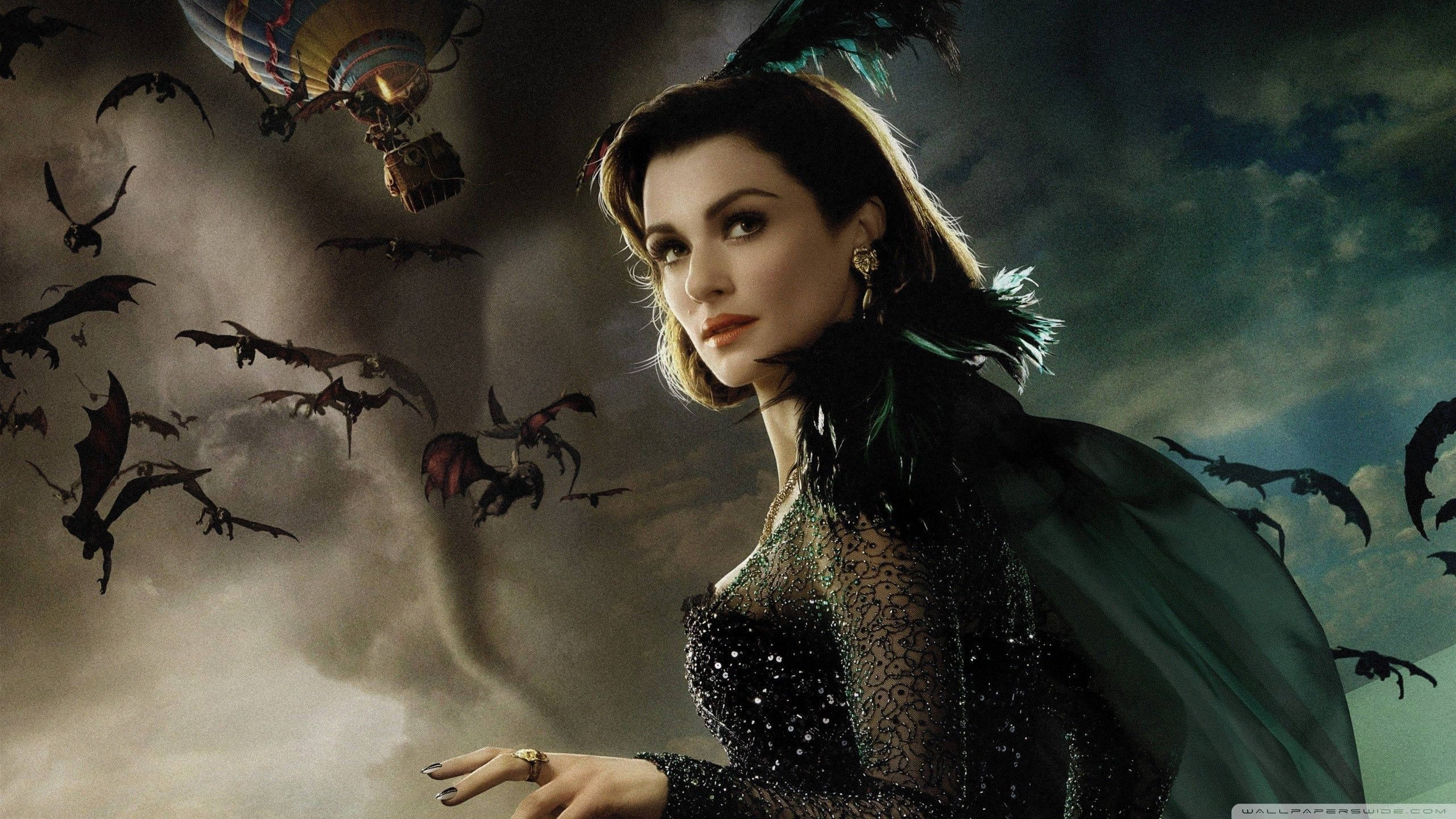 Rachel Weisz 桌布 壁紙 — 一位身穿深色蕾絲長裙和綠色羽毛披肩的女性站在暴風雨的天空下，蝙蝠圍繞著熱氣球飛舞。