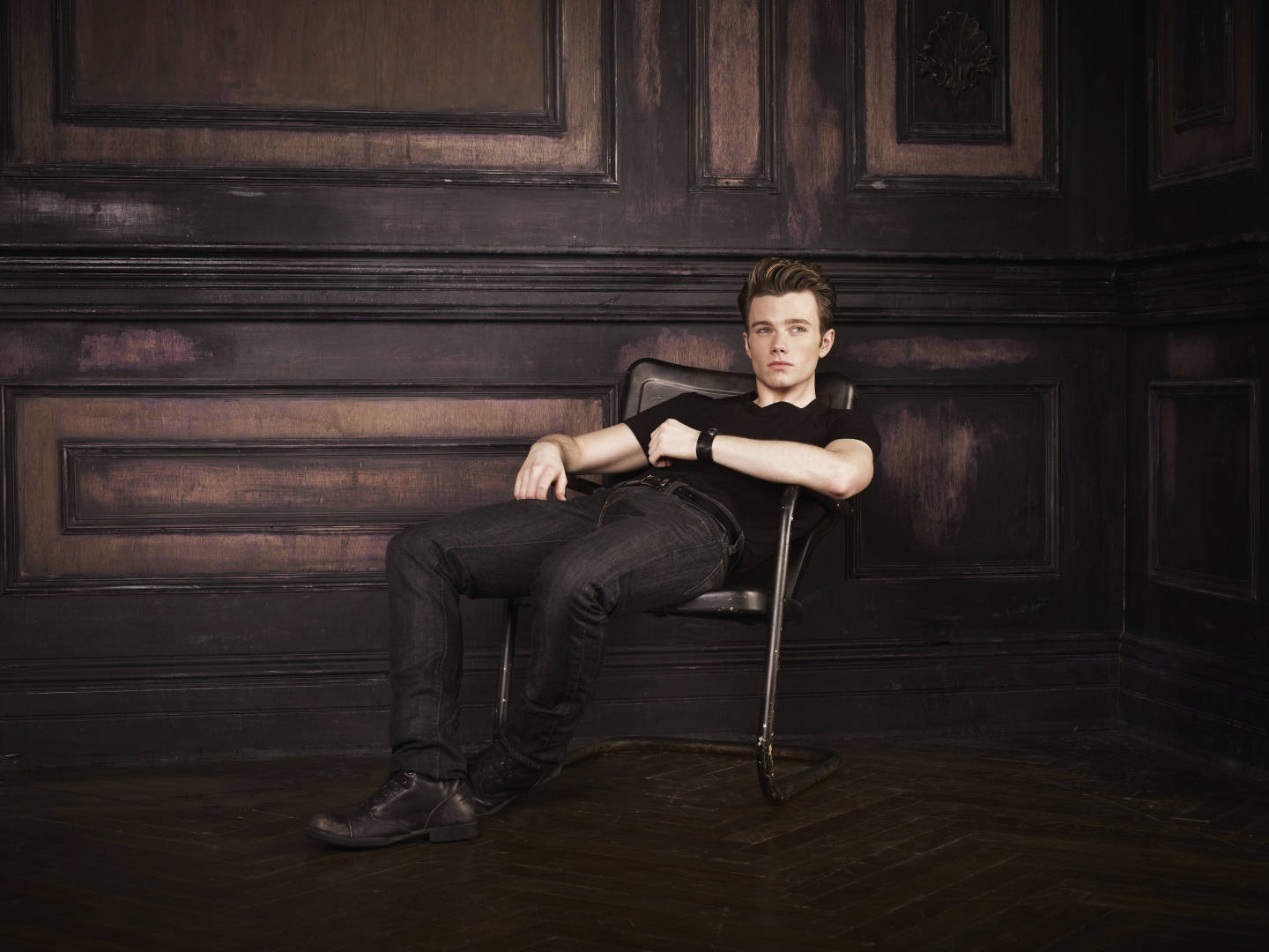 Chris Colfer 桌布 壁紙 — 一位身穿深色衣服的年輕男子，在光線昏暗的房間裡，靠著華麗的鑲板木牆，放鬆地坐在椅子上。