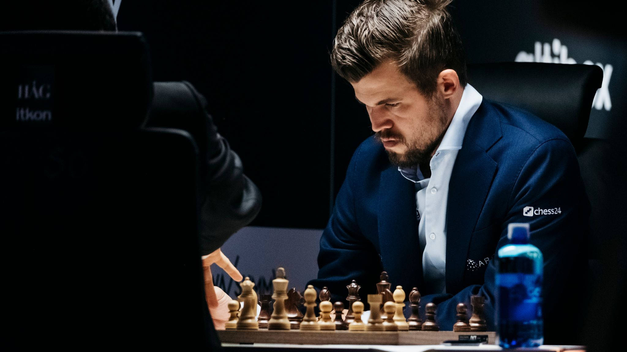 Magnus 桌布 壁紙 — 一位身穿深色西裝、專注的西洋棋選手在昏暗的比賽環境中俯身於棋盤之上。